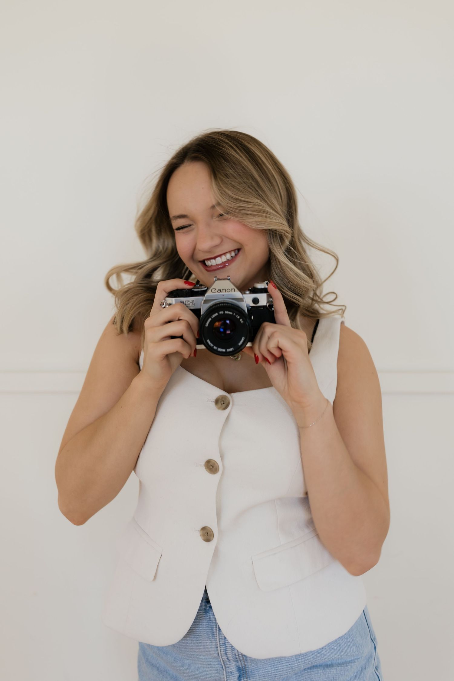 Person in white sleeveless top and jeans holds vintage camera while smiling and posing against white background.