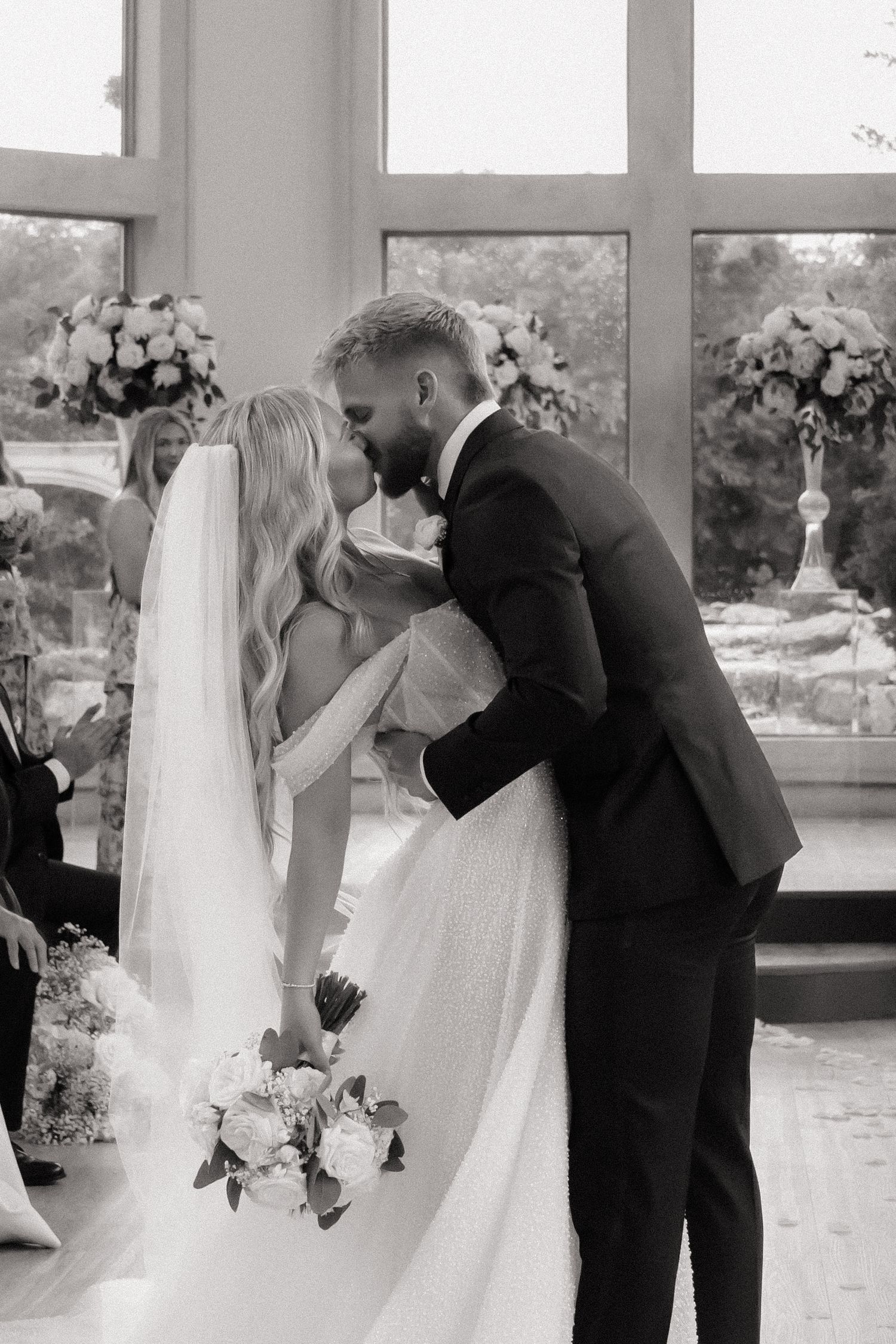 A romantic wedding ceremony kiss captured in black and white by large windows with floral arrangements.