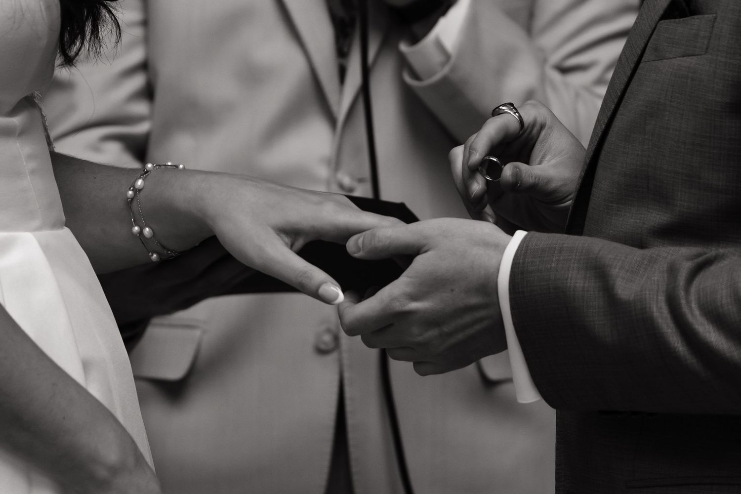 Black and white photo of a couple exchanging wedding rings during a ceremony, with a bracelet visible on the bride's wrist.