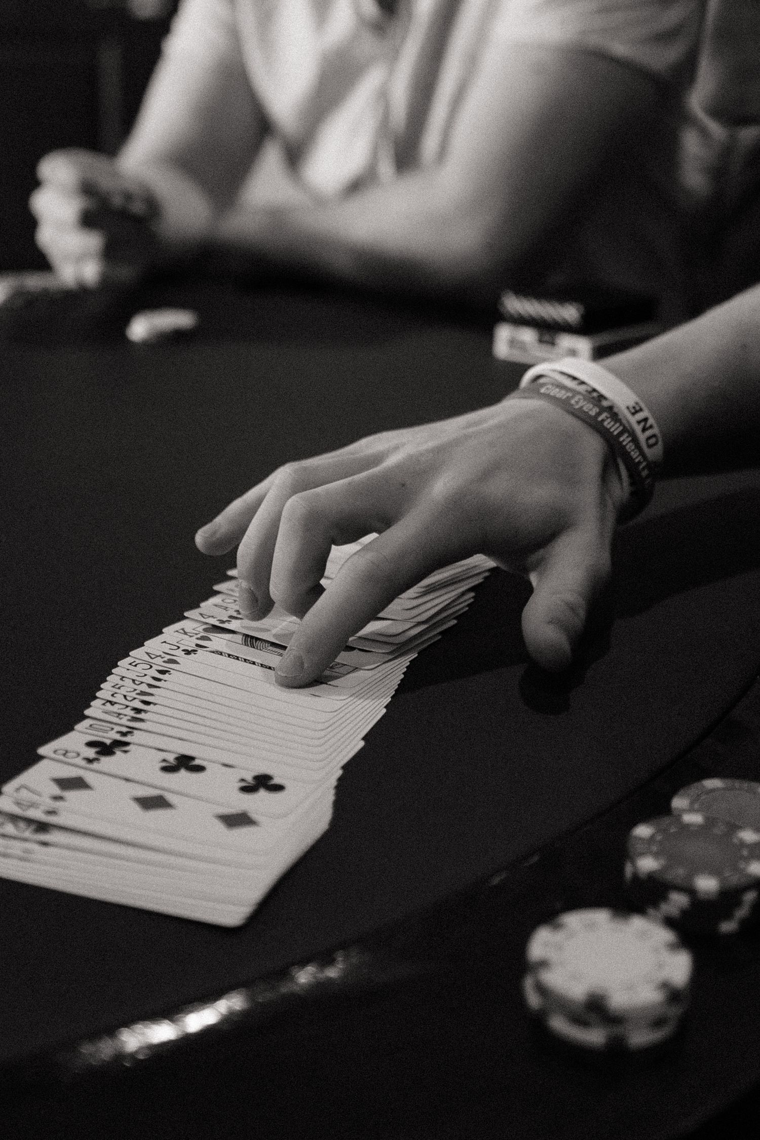 Black and white close-up of hands dealing playing cards on a dark table with poker chips.