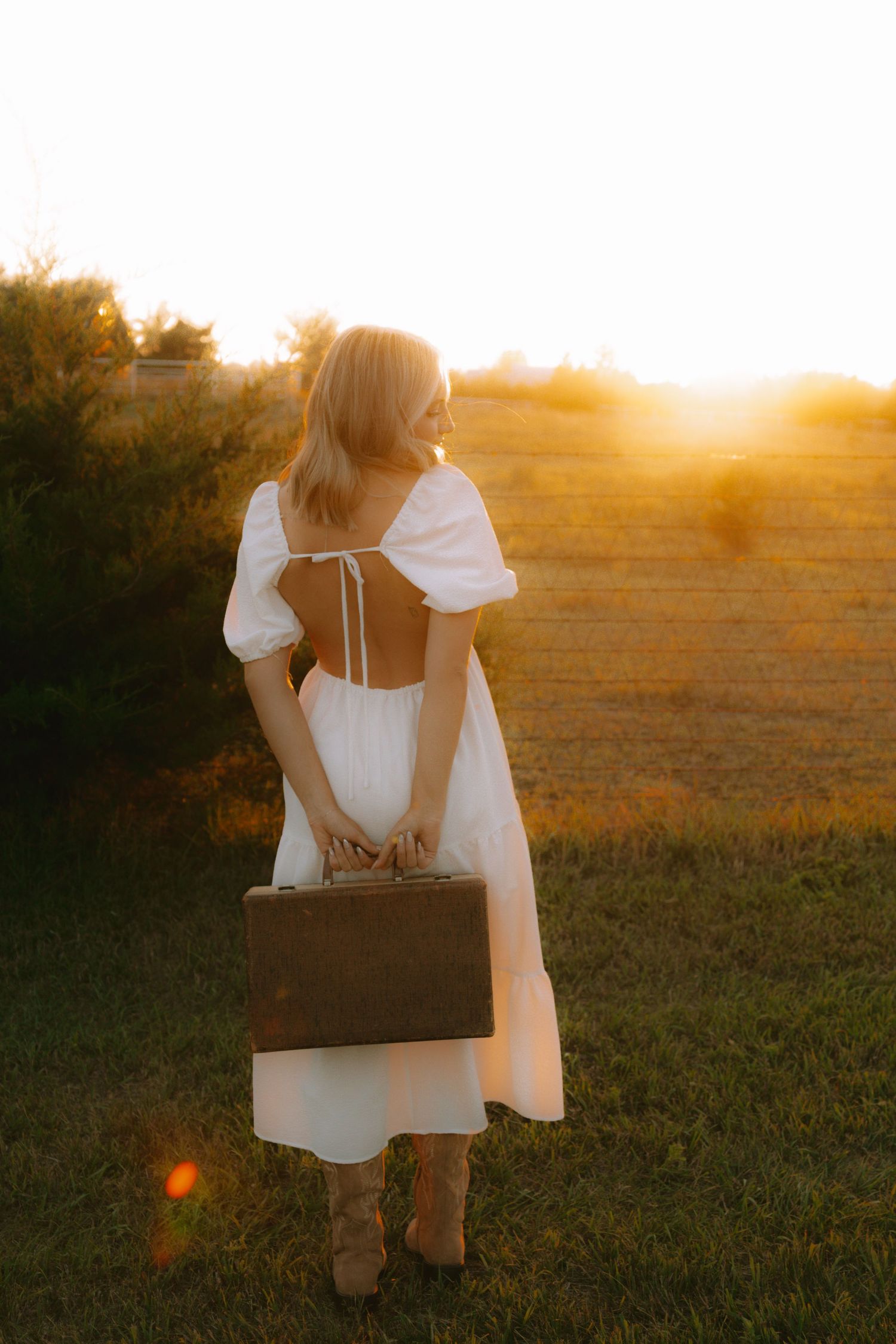 A figure in a white dress holds a vintage suitcase while standing in a golden sunlit field at sunset.