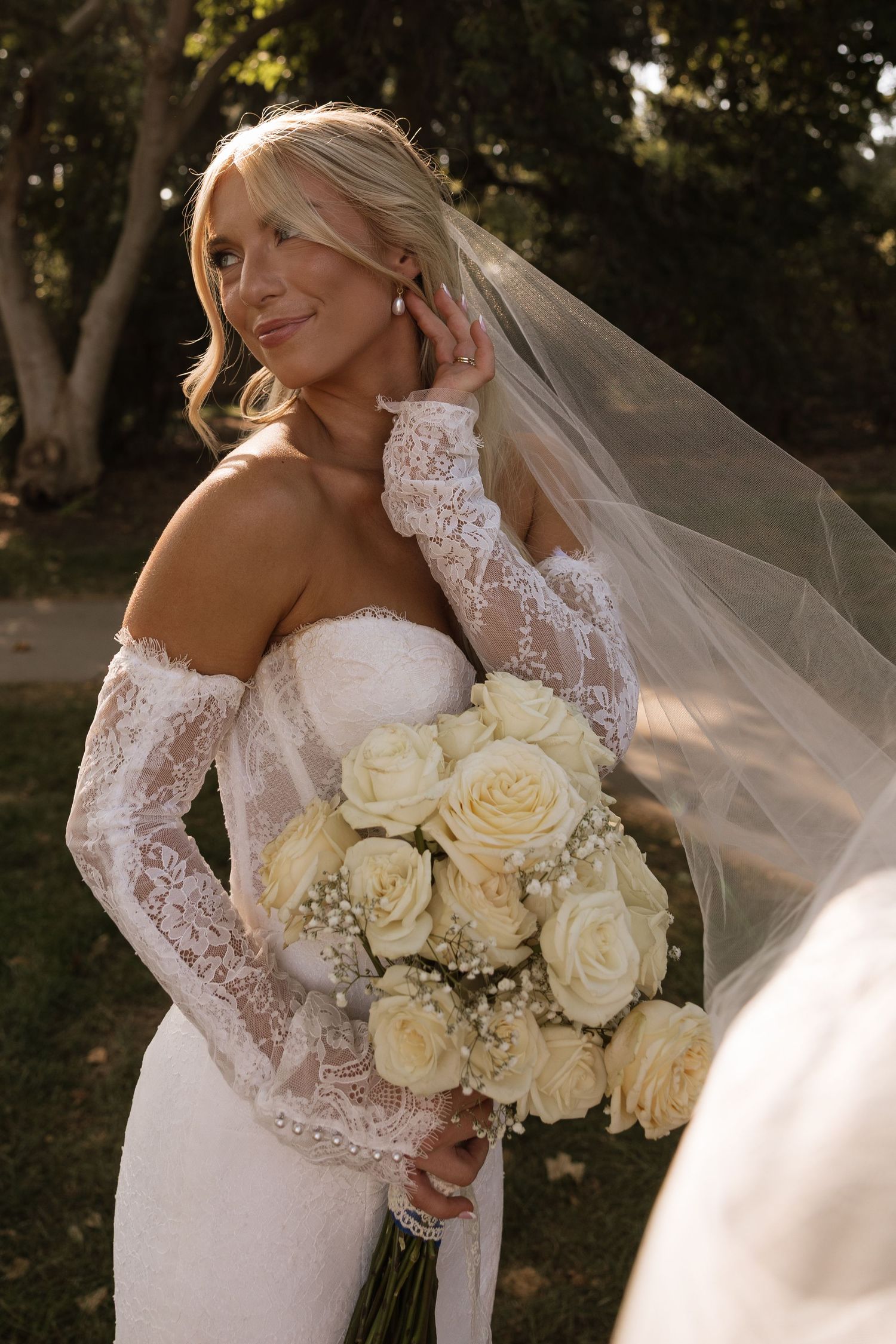 Blonde bride in strapless lace gown with detachable sleeves and veil holds white rose bouquet outdoors.