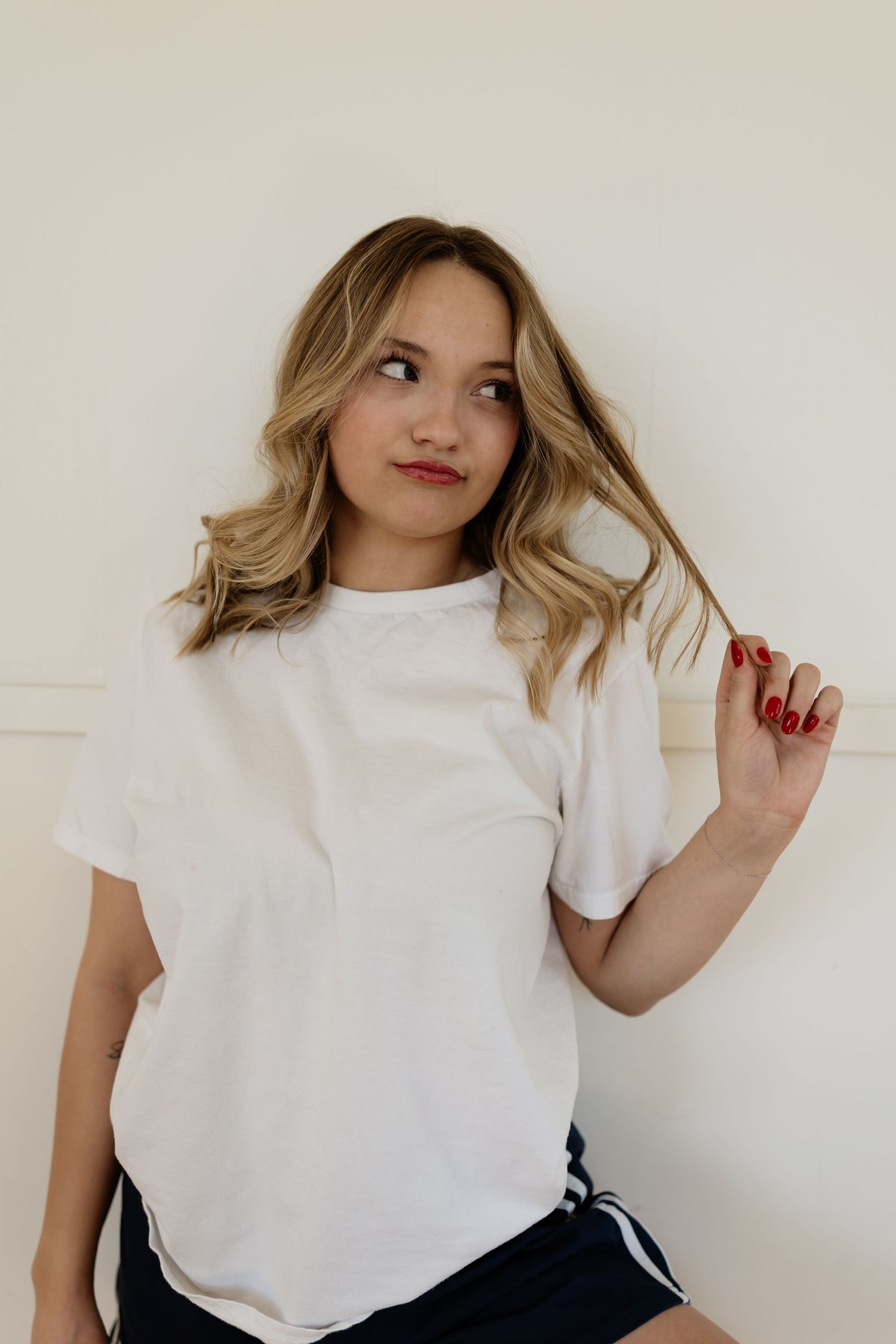 Person in white t-shirt poses against white wall while playing with blonde wavy hair.