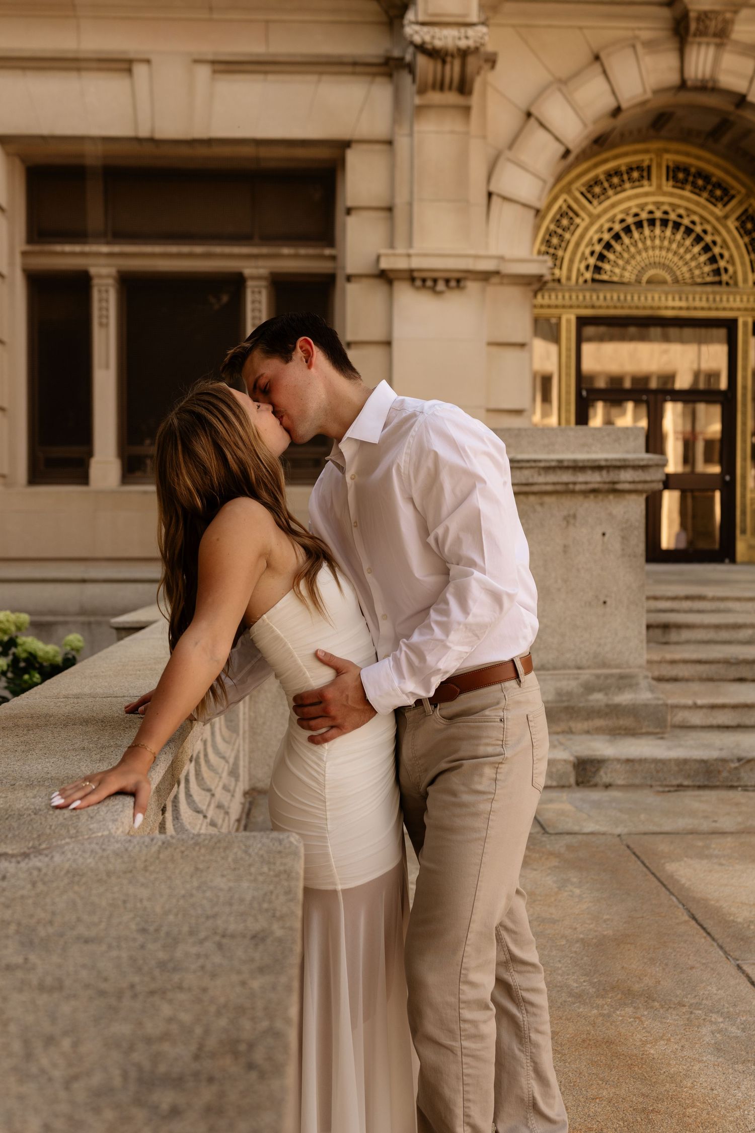 Couple sharing a romantic kiss in elegant neutral outfits on stone steps outside a classic building.