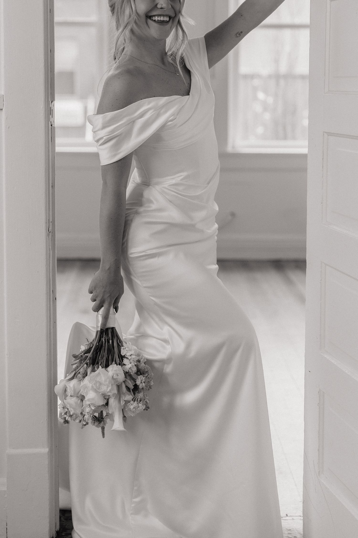 Candid black and white photo of a bride in a sleek white wedding dress holding flowers.