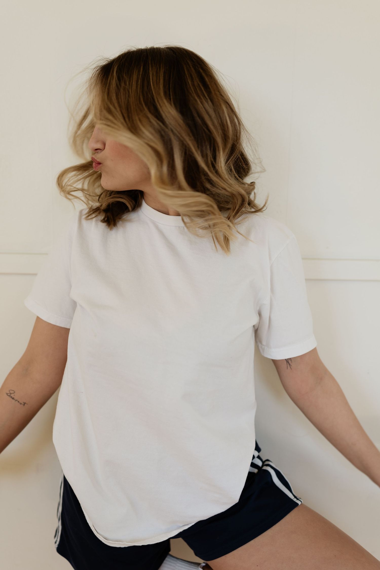 A person wearing a white t-shirt and dark shorts sits on a white surface in a minimalist setting.