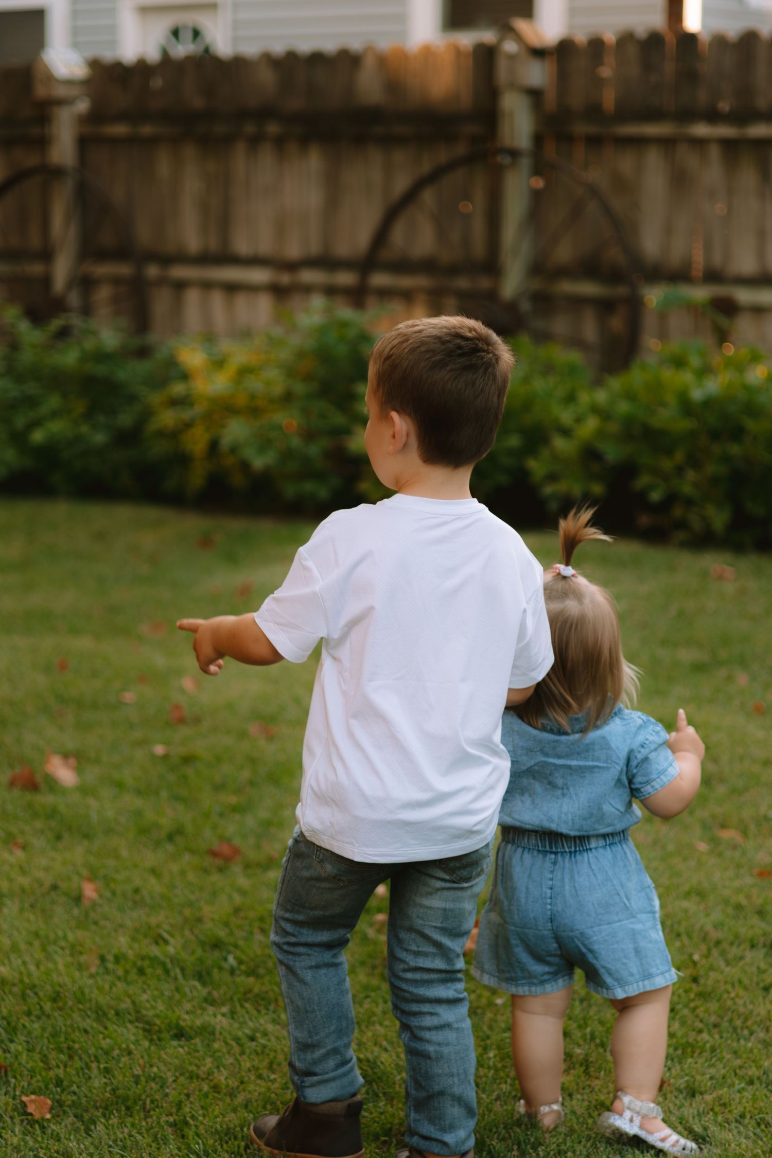 Two children point and look at something in a backyard garden with a wooden fence.