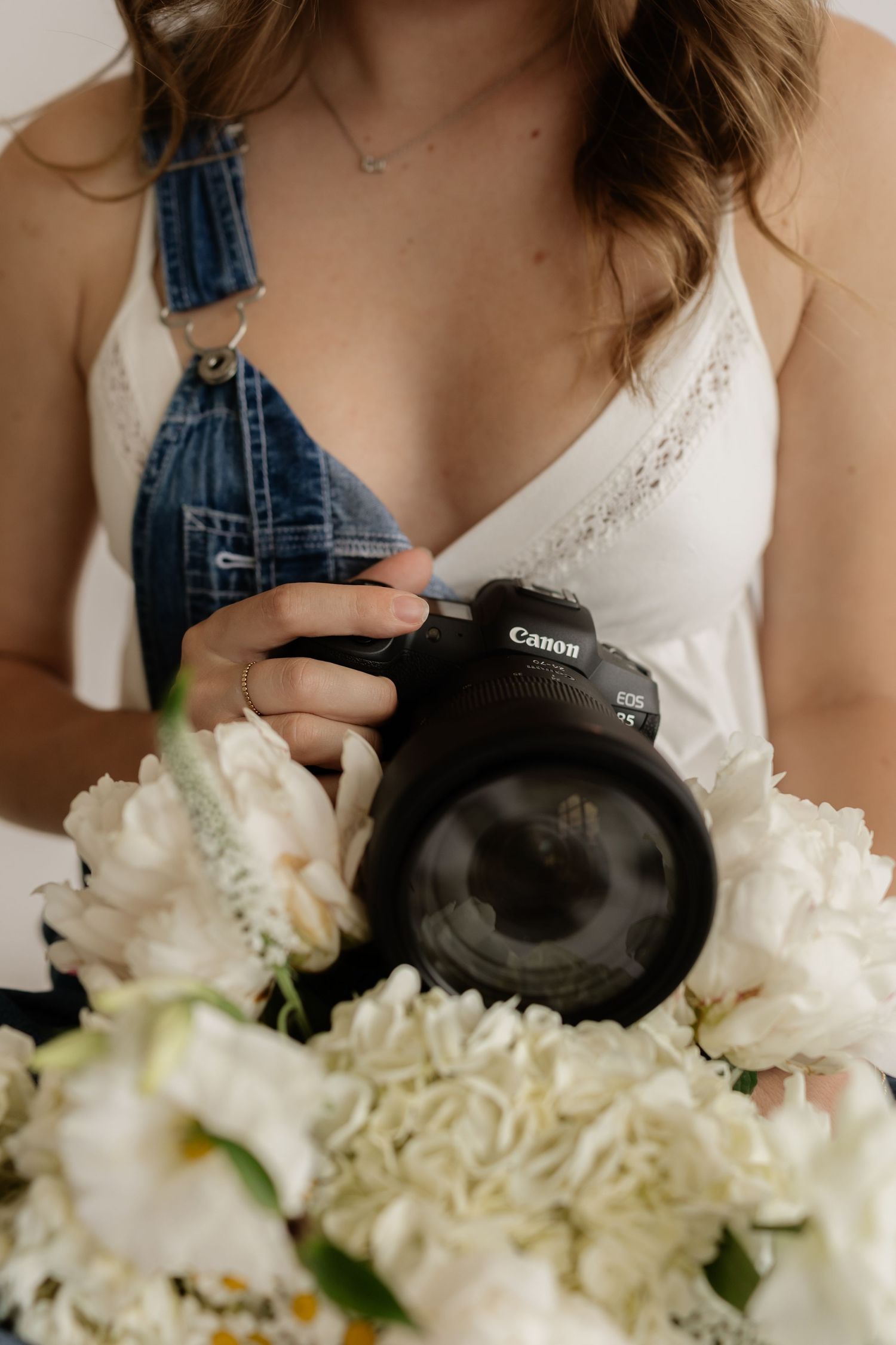 Woman in denim overalls holding a Canon DSLR camera over white peony flowers.