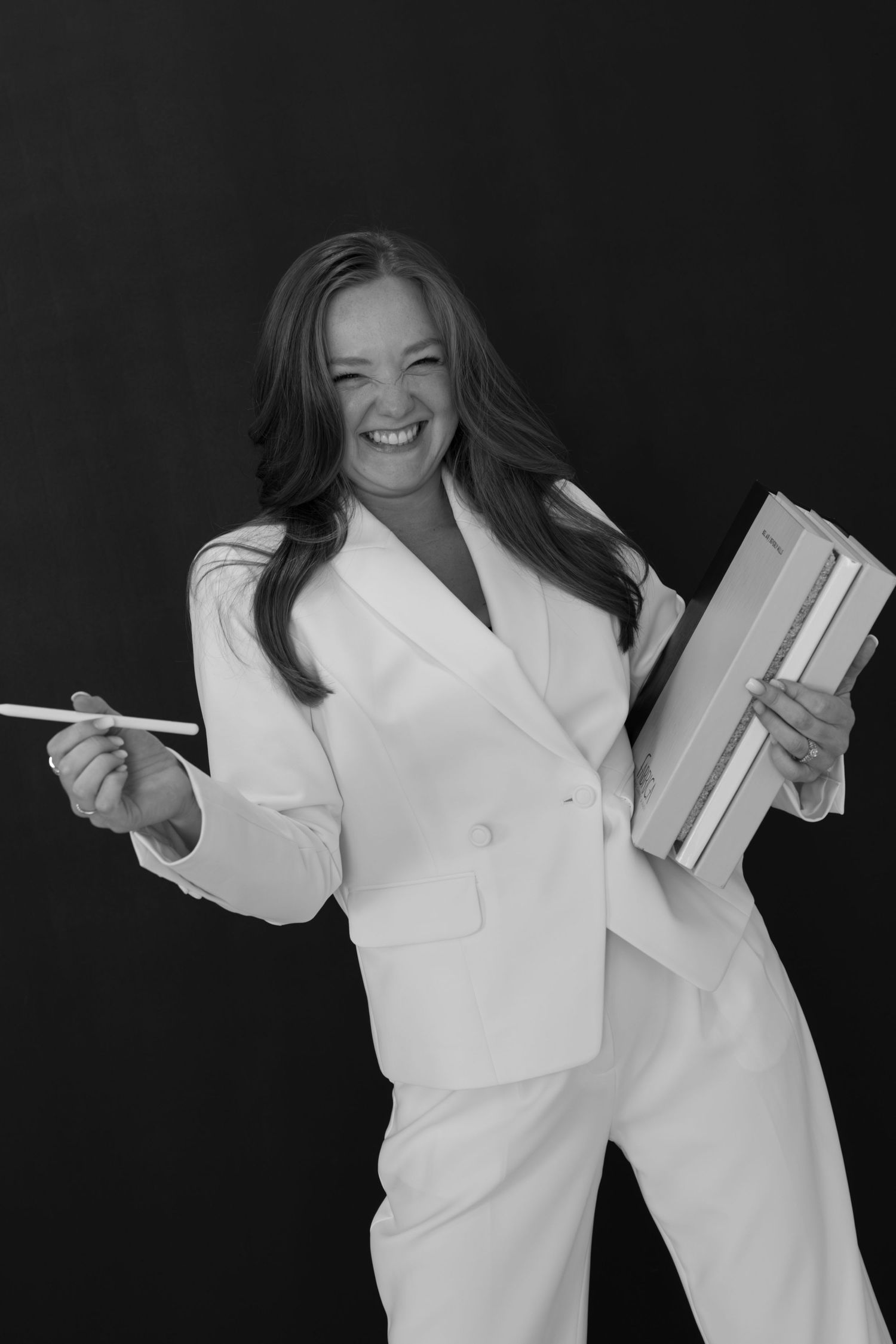 Professional in white suit holds books and pen while expressing joy against dark background.