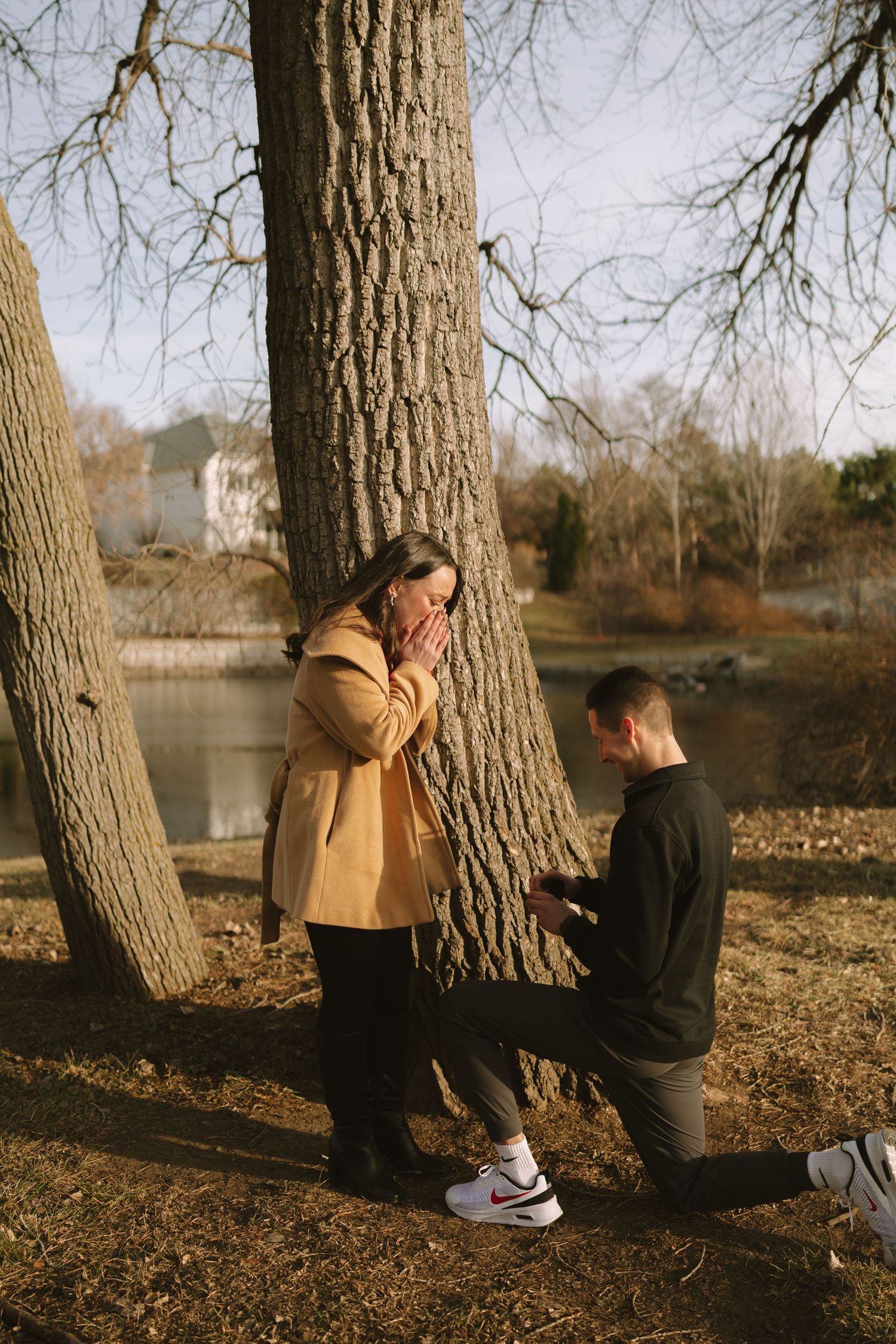 A surprise proposal moment captured between two people by a large tree near a lake.