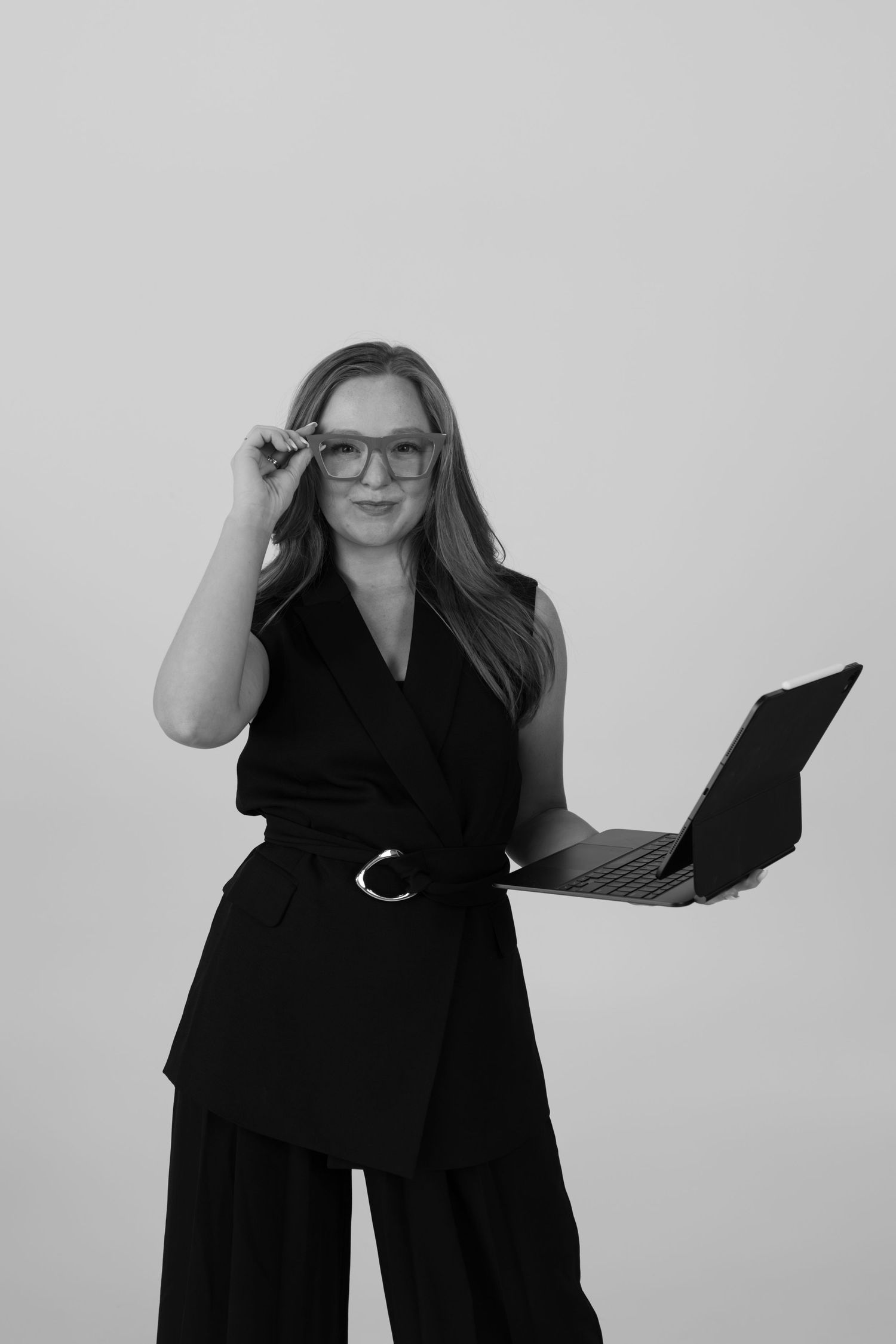 Professional in black attire adjusts glasses while holding laptop against minimalist background.
