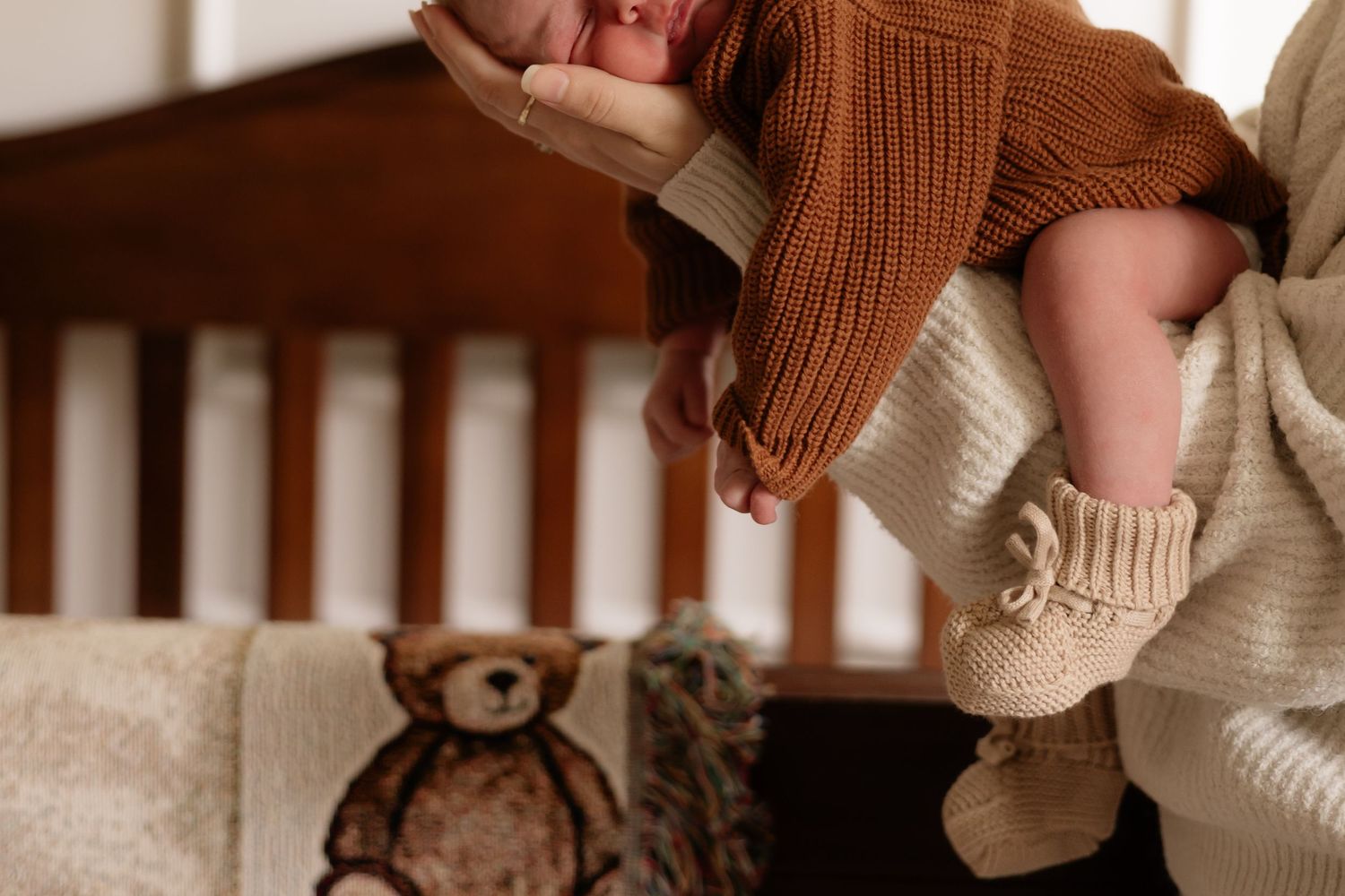 Parent holding newborn baby in brown knit outfit near a wooden crib with a teddy bear blanket.