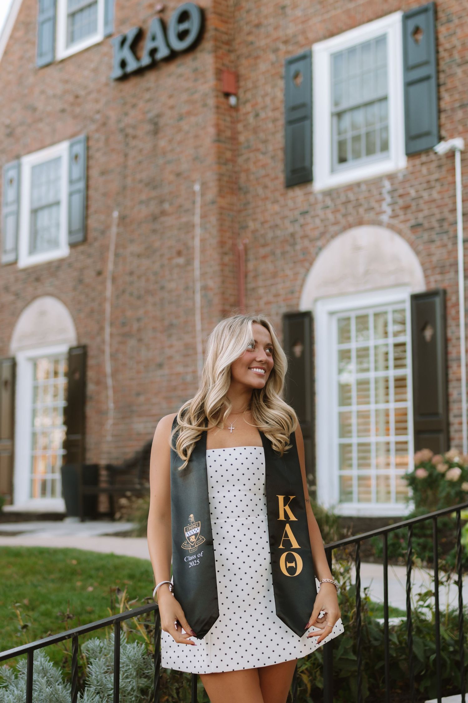 Graduate in polka dot dress and sorority stole poses outside brick Kappa Theta house.