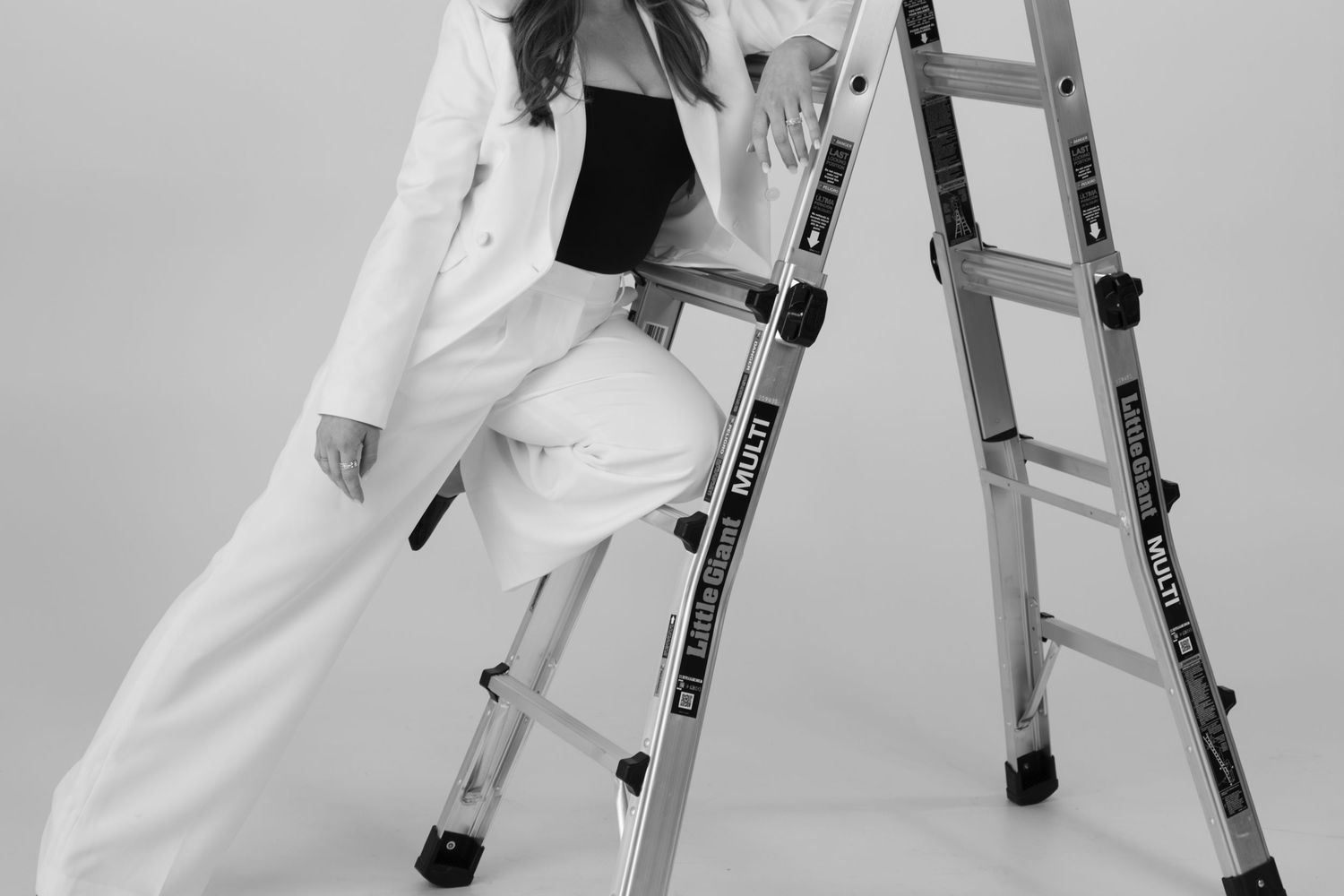 Person in crisp white suit poses confidently on an aluminum ladder against gray backdrop.