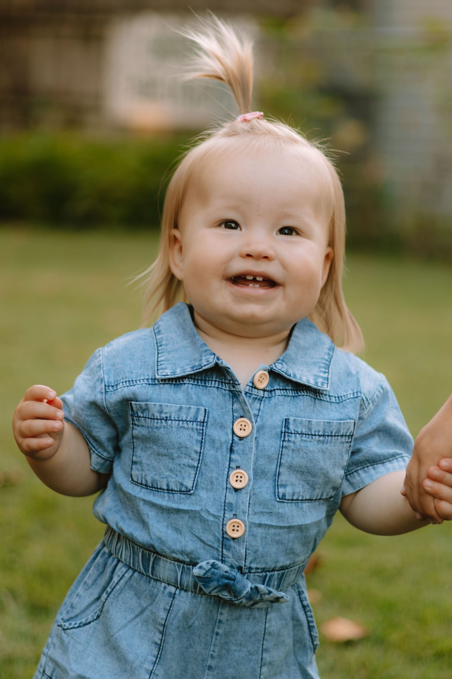A toddler in a denim dress smiles while playing outdoors.