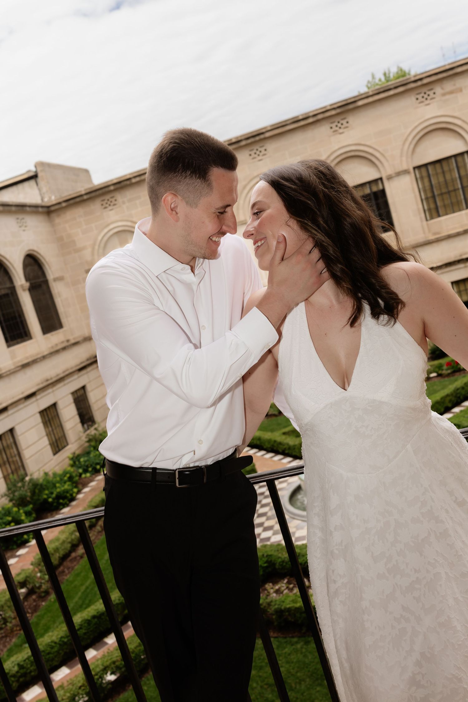 A groom tenderly holds his bride's face as they share an intimate moment on a balcony outside a historic stone building.