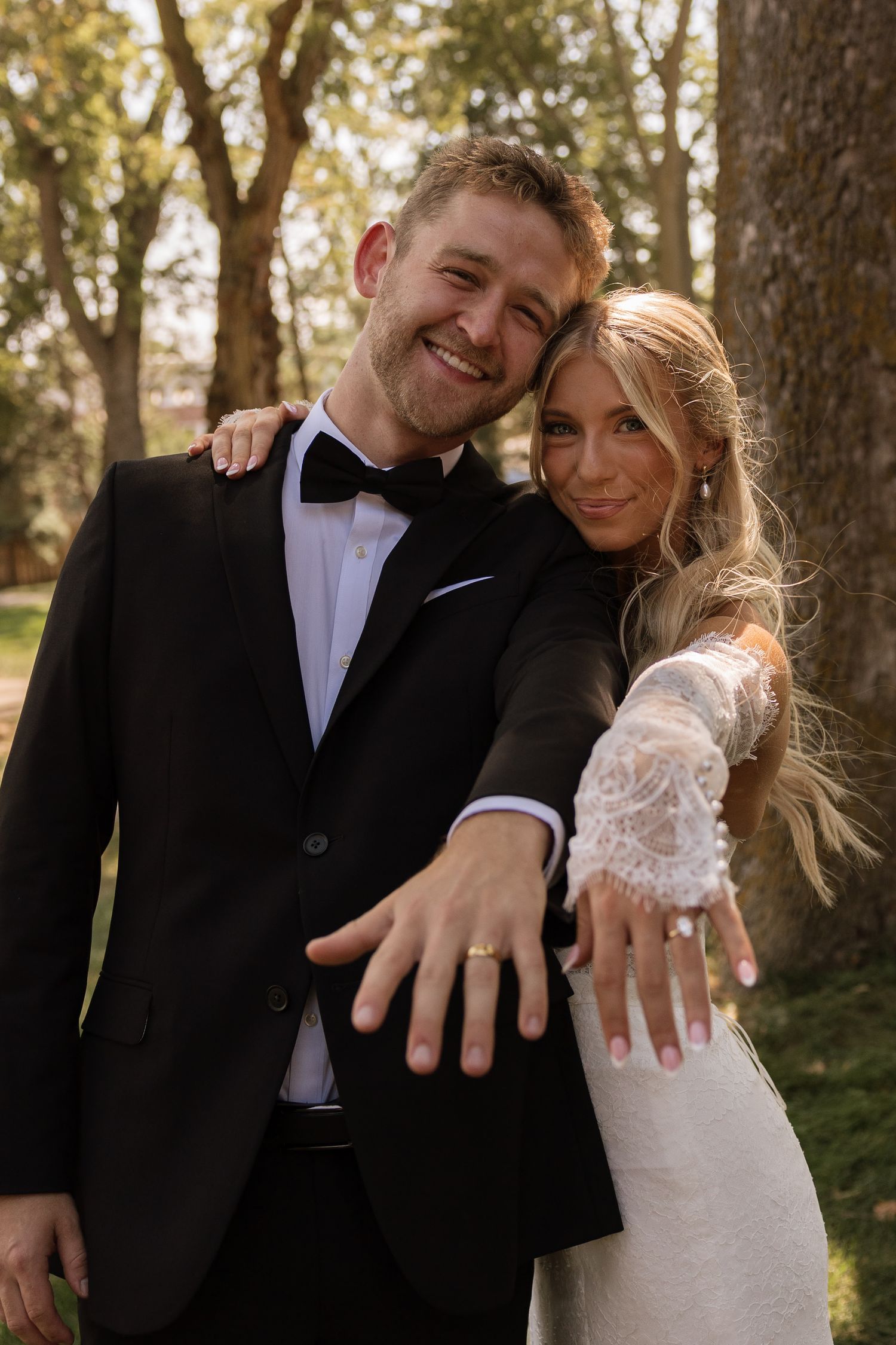 Happy newlyweds in formal attire showing off wedding rings outdoors, bride in lace gown and groom in black tuxedo.