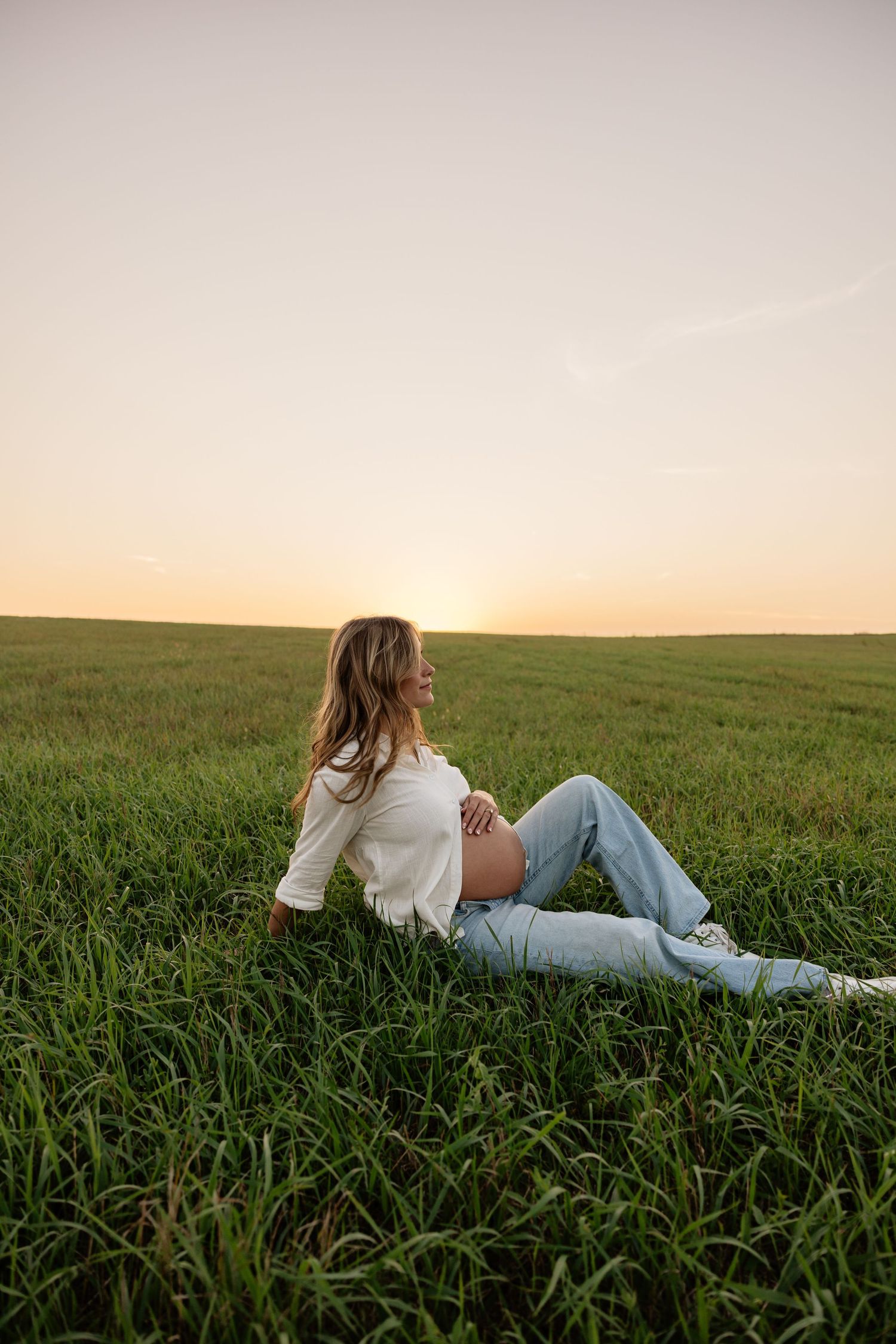 Pregnant woman in white top and jeans relaxing in green field at sunset, smiling peacefully.