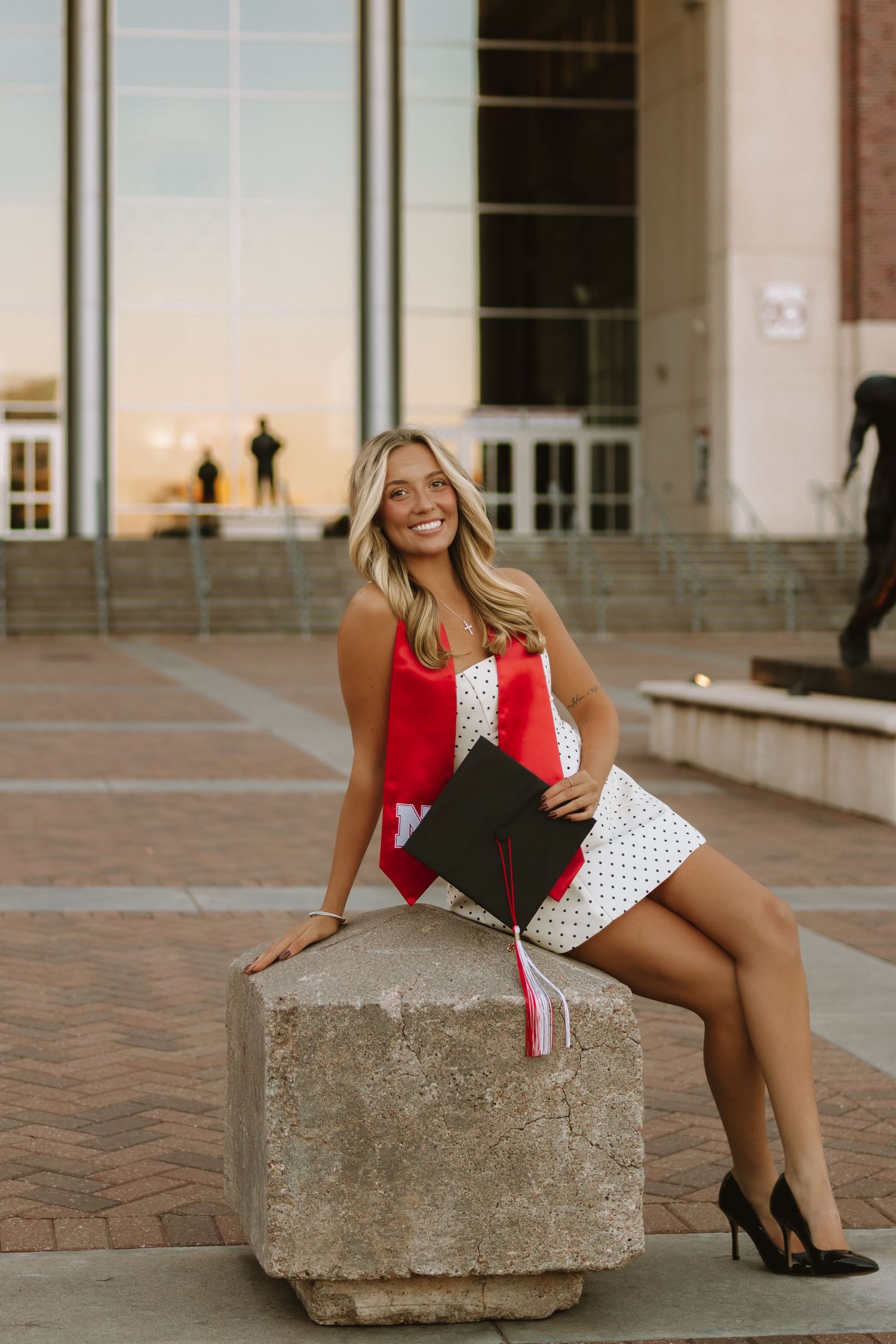 Graduate in white polka dot dress with red sash poses on concrete block in front of building.