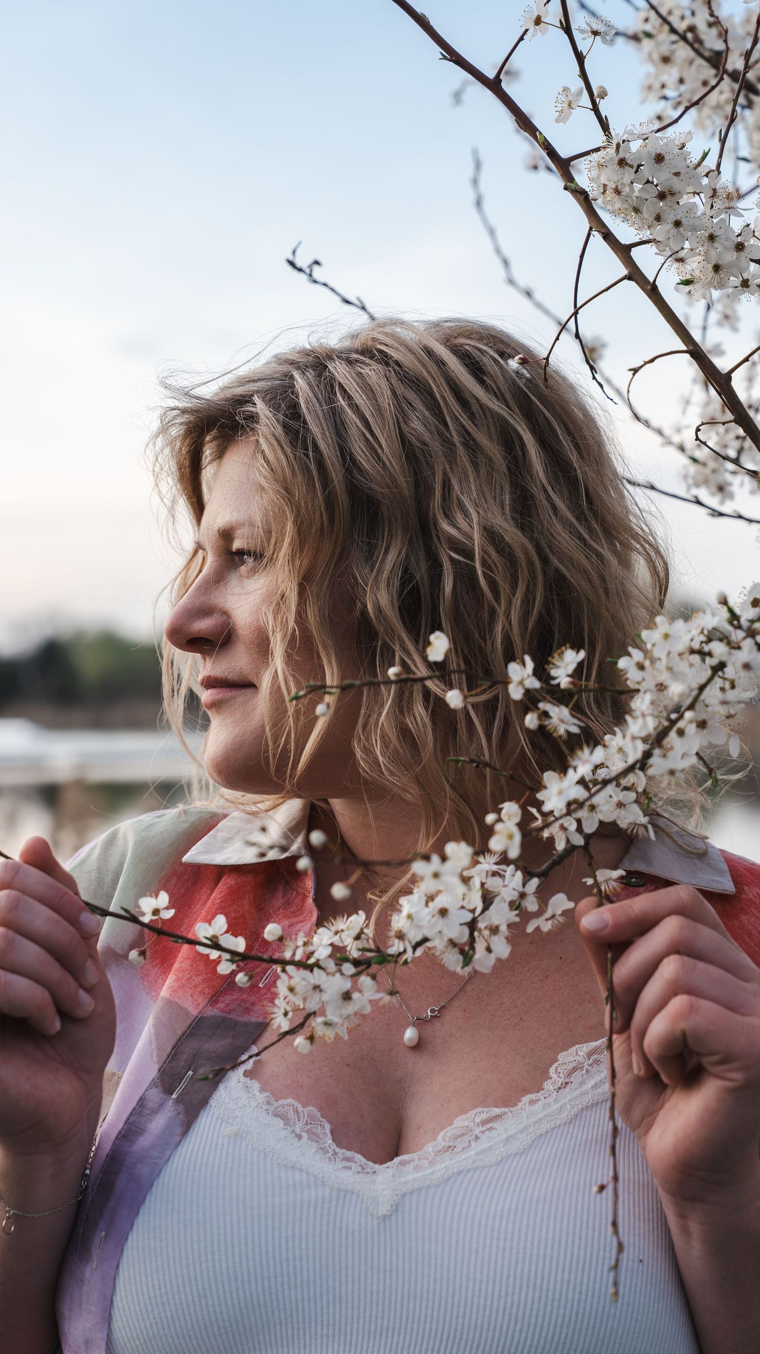 Woman with wavy blonde hair holding cherry blossom branches outdoors in spring.