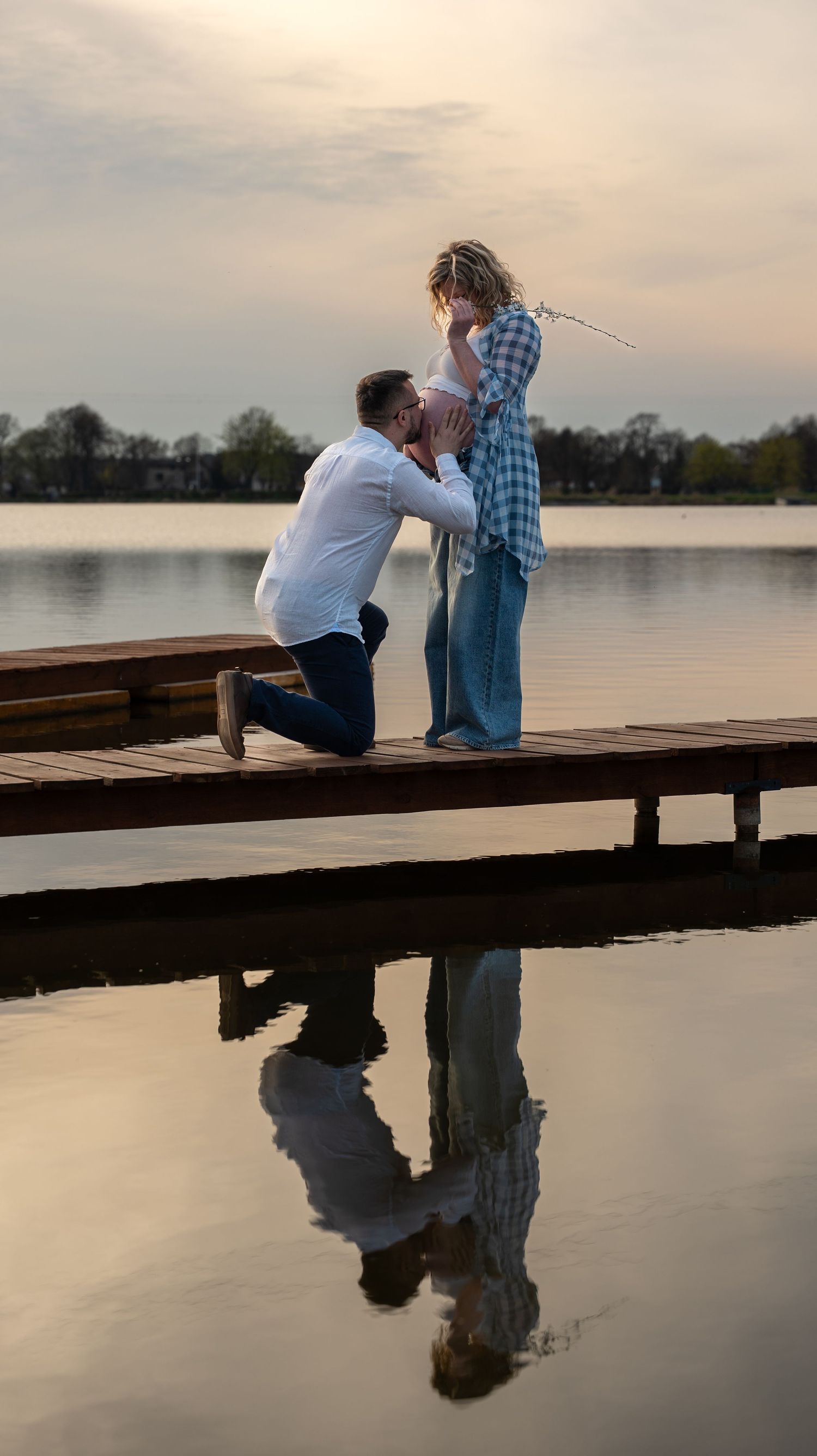 Man kneeling on a wooden dock proposing to a pregnant woman at sunset by a calm reflective lake.