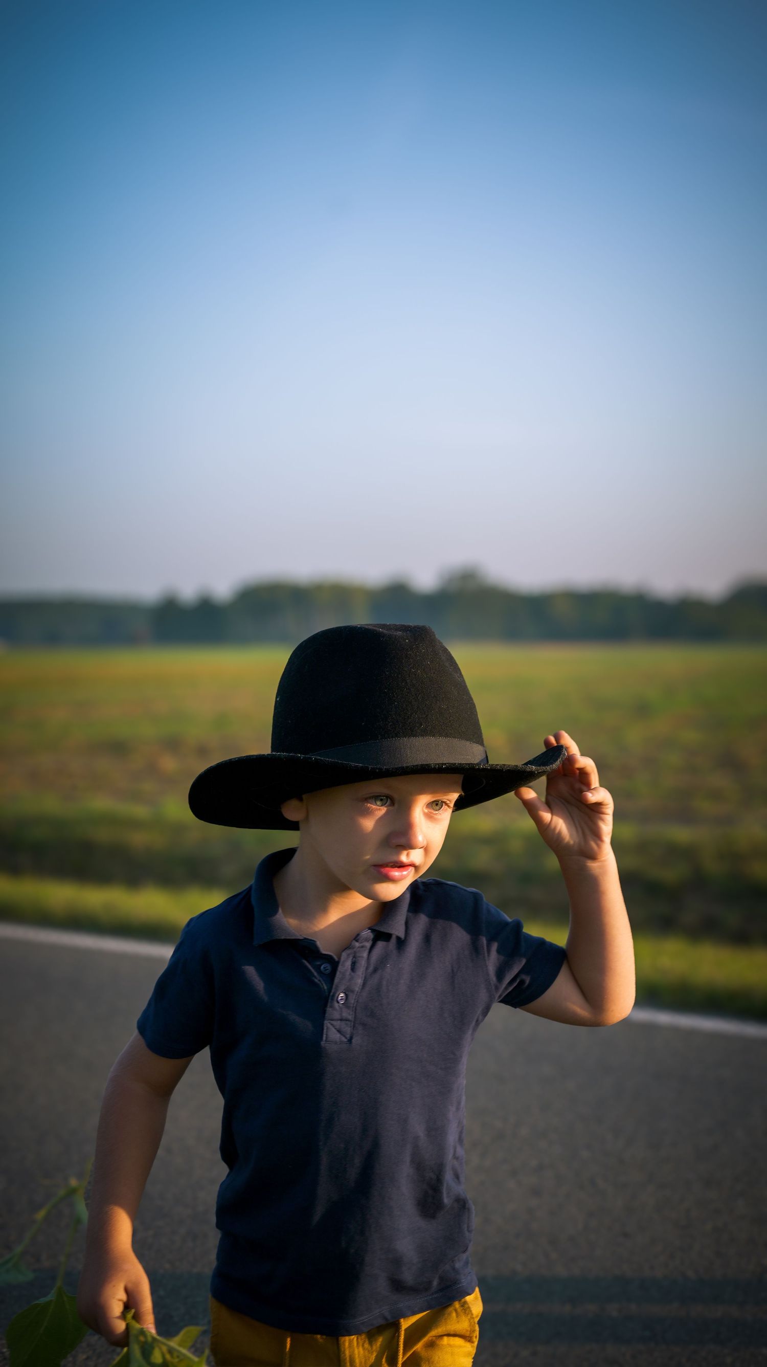 Child in navy shirt and black hat standing on a road with blue sky and fields in background.
