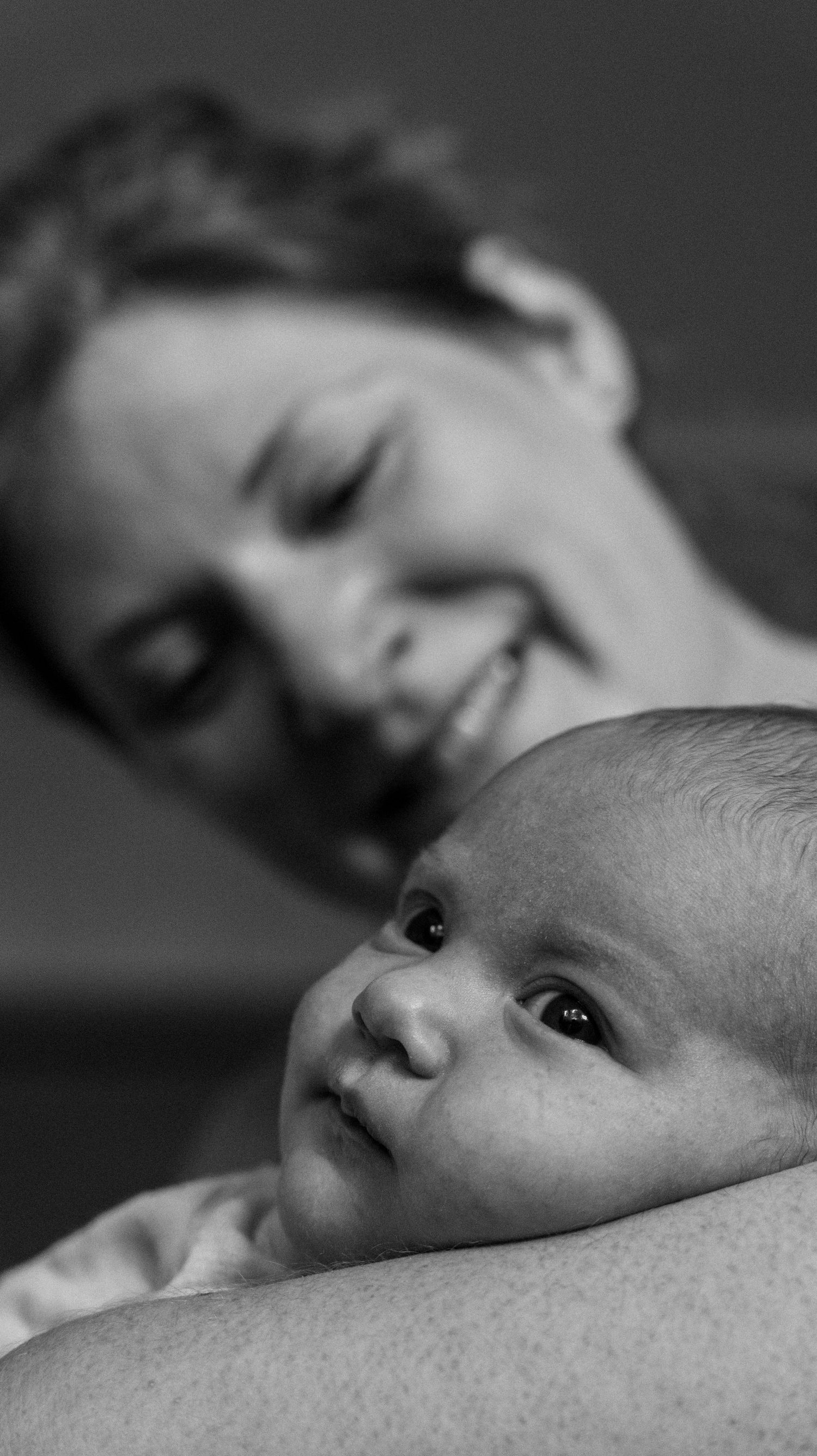 Black and white portrait of a smiling mother gazing lovingly at her newborn baby looking up with wide eyes.