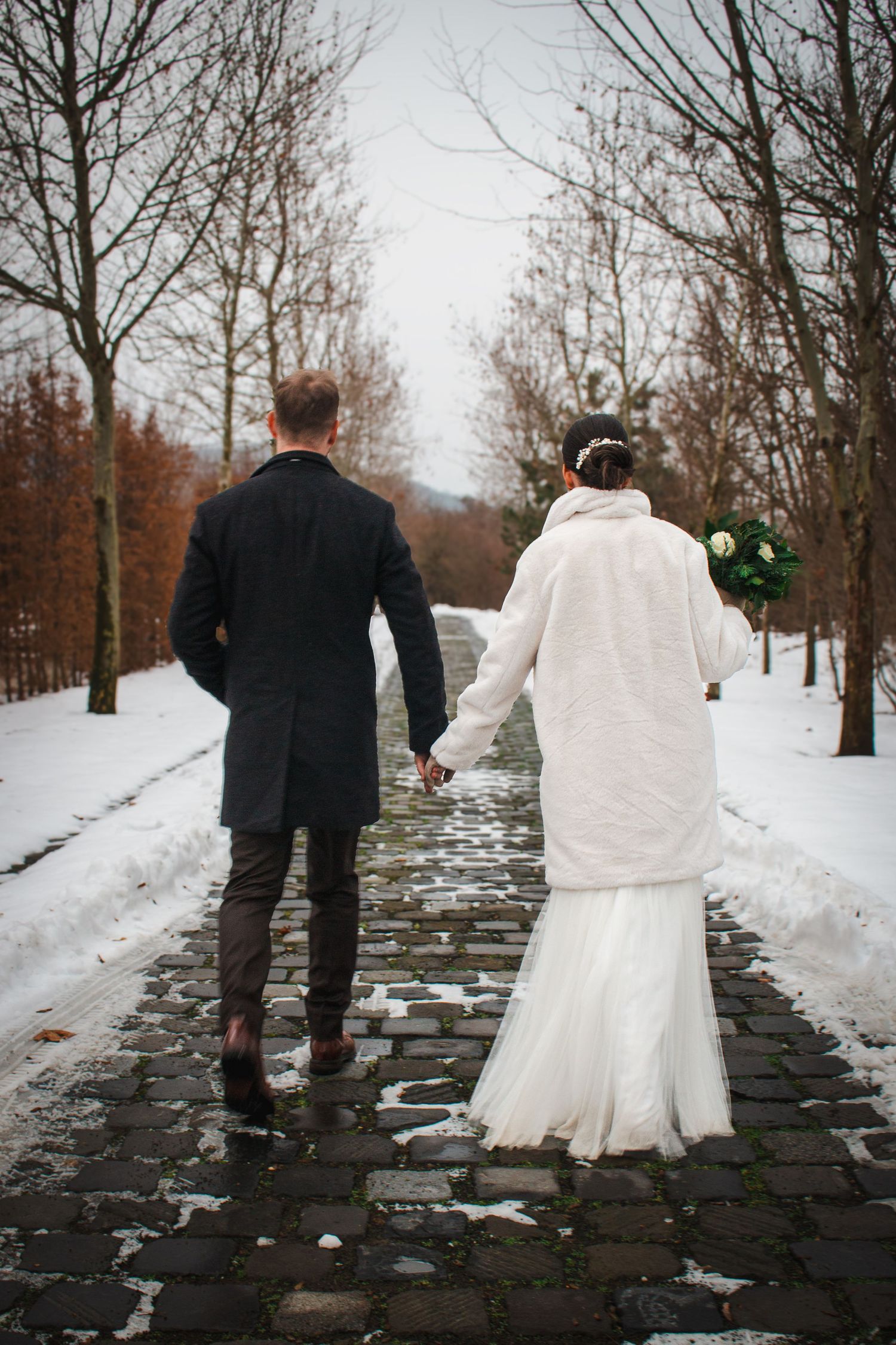 Wedding couple walking away on snowy path lined with bare winter trees.