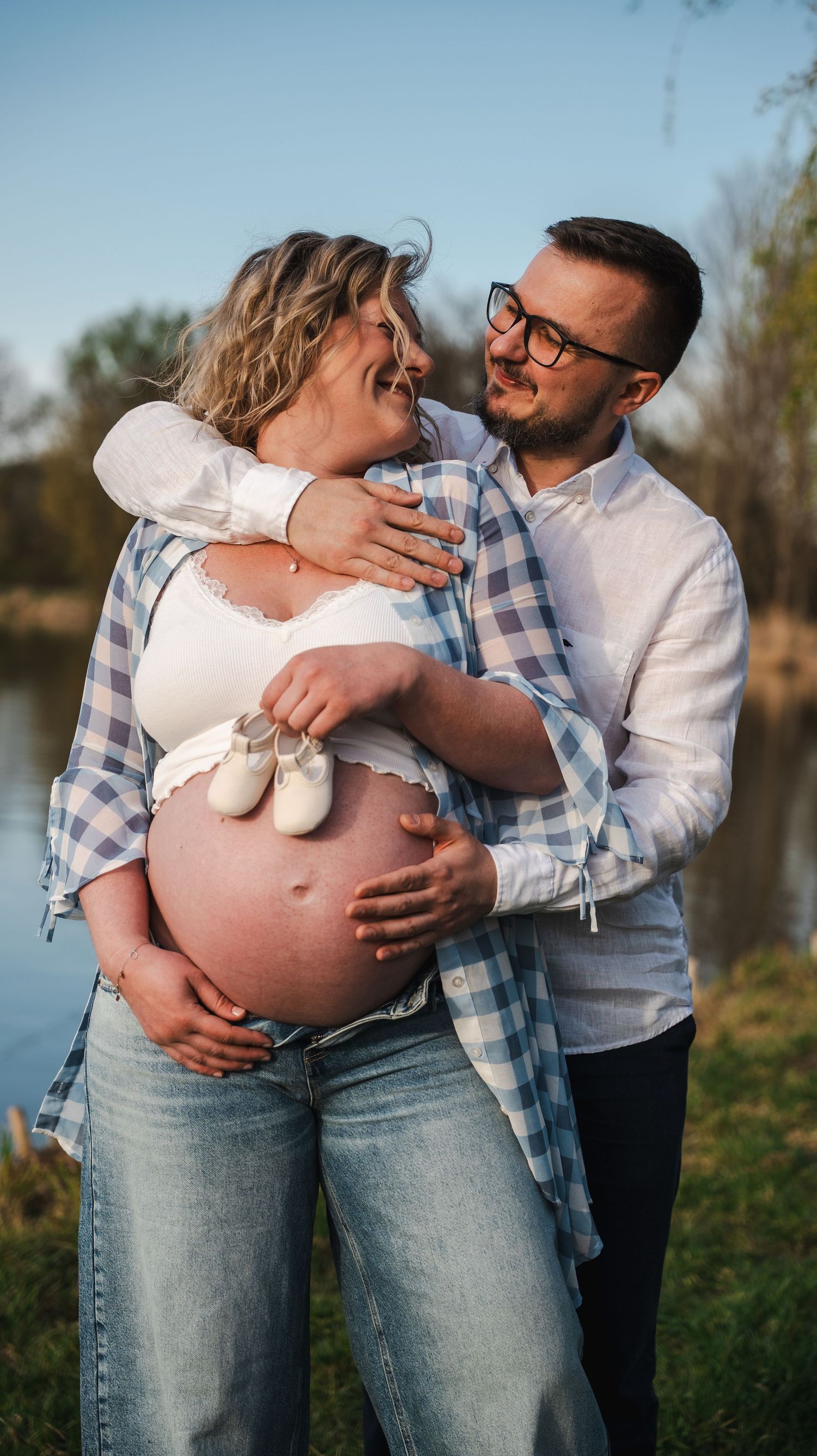 Happy pregnant couple embracing outdoors by lake, holding tiny baby shoes on baby bump.