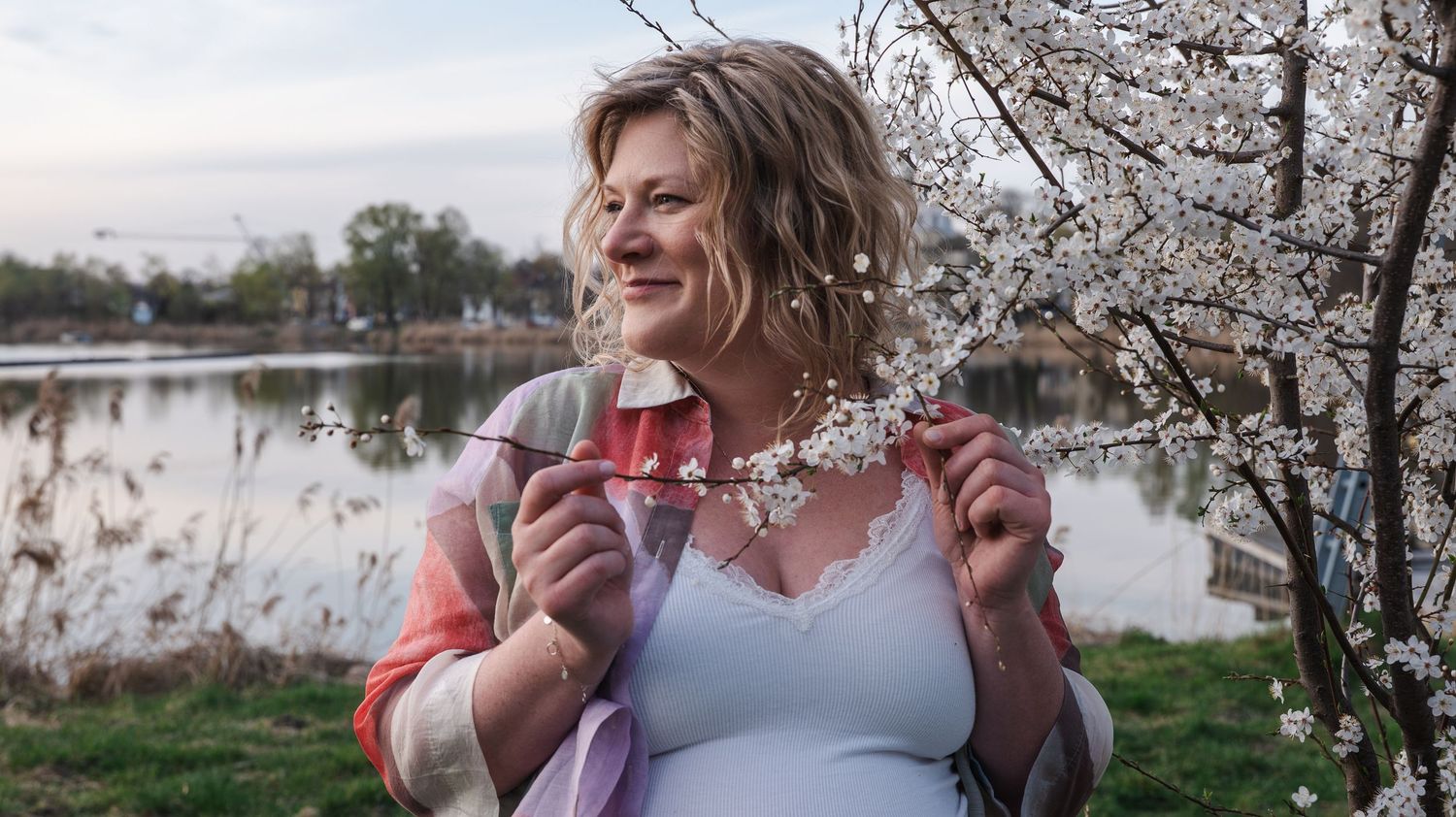 A smiling woman stands near a blooming cherry tree by a lake, wearing a white top and pink cardigan in spring.