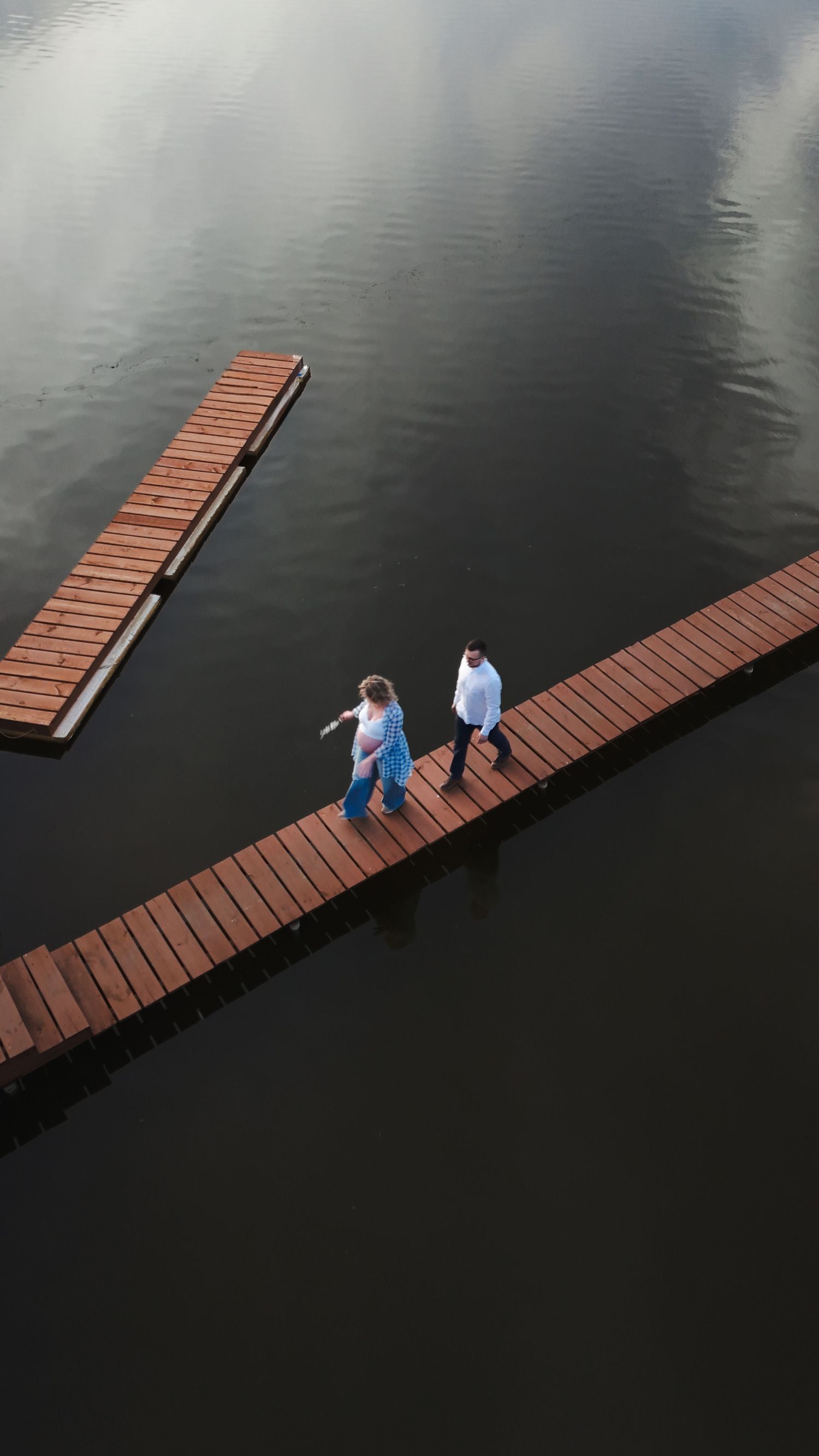 Aerial view of two people walking on a wooden floating dock over calm dark water.