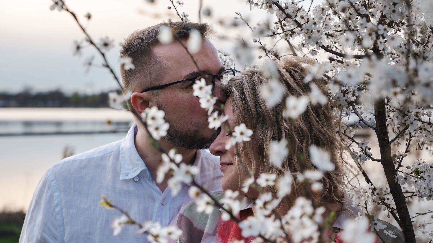 Couple sharing a romantic moment among white cherry blossoms with a serene lake backdrop at sunset.