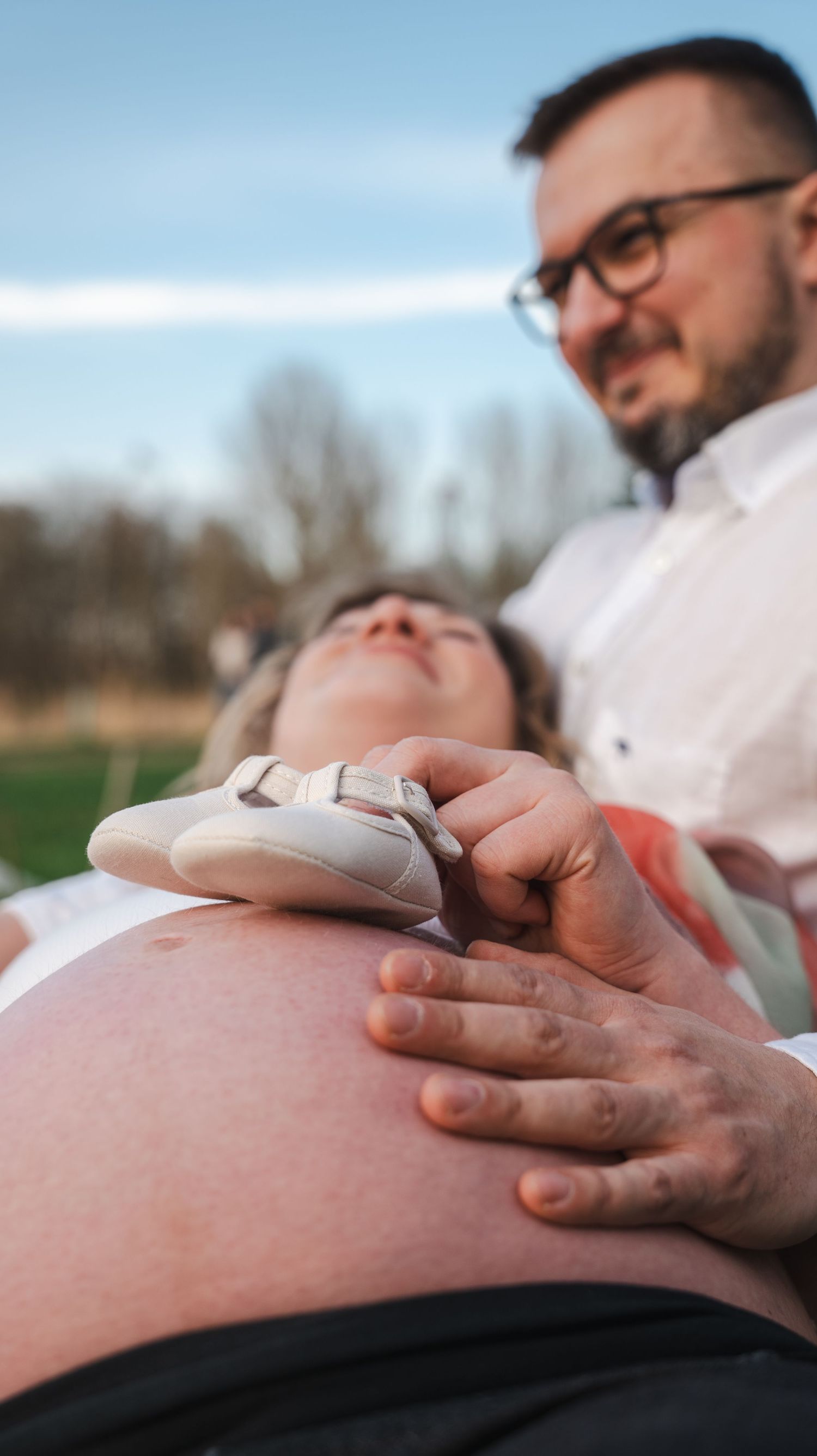 Pregnant woman lying down as her partner holds tiny baby shoes on her bare bump outdoors.