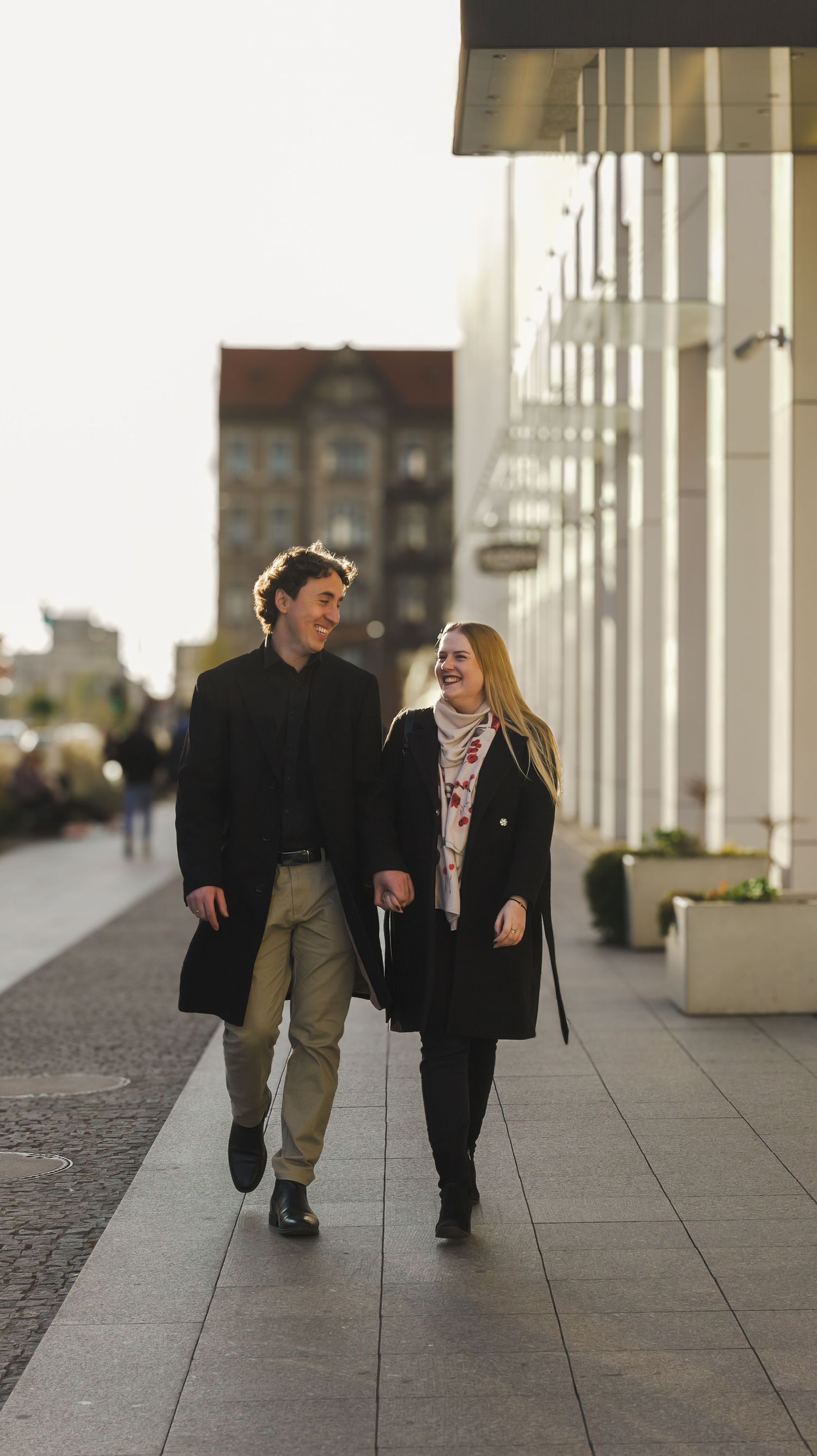 Two people walking together and laughing along a colonnade with white pillars.