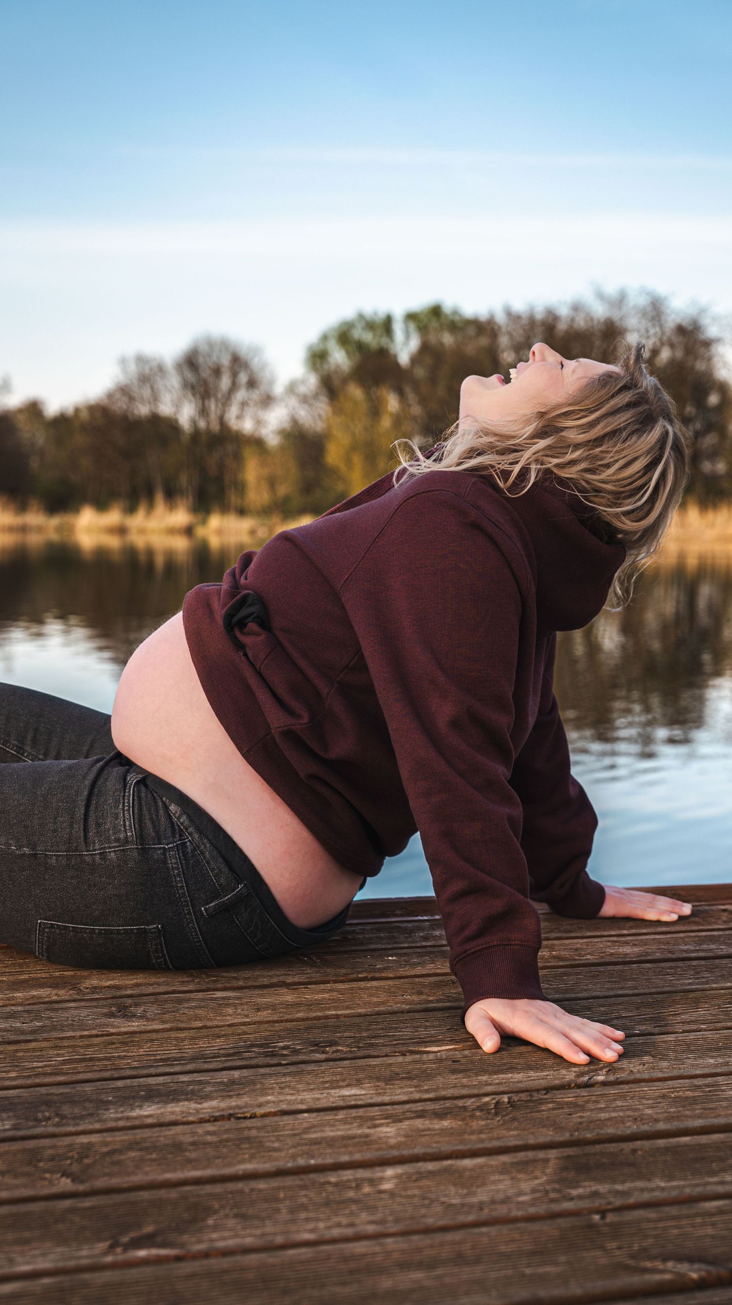 Pregnant woman in burgundy hoodie and jeans sitting on wooden dock by lake, head tilted back.