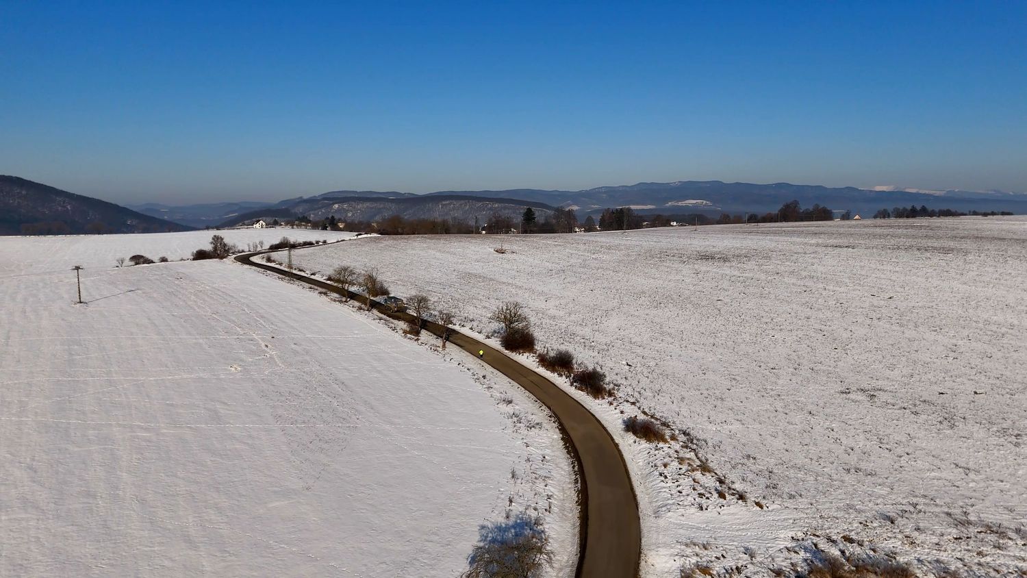 A winding road curves through a snowy winter landscape with mountains visible in the distance under clear blue skies.