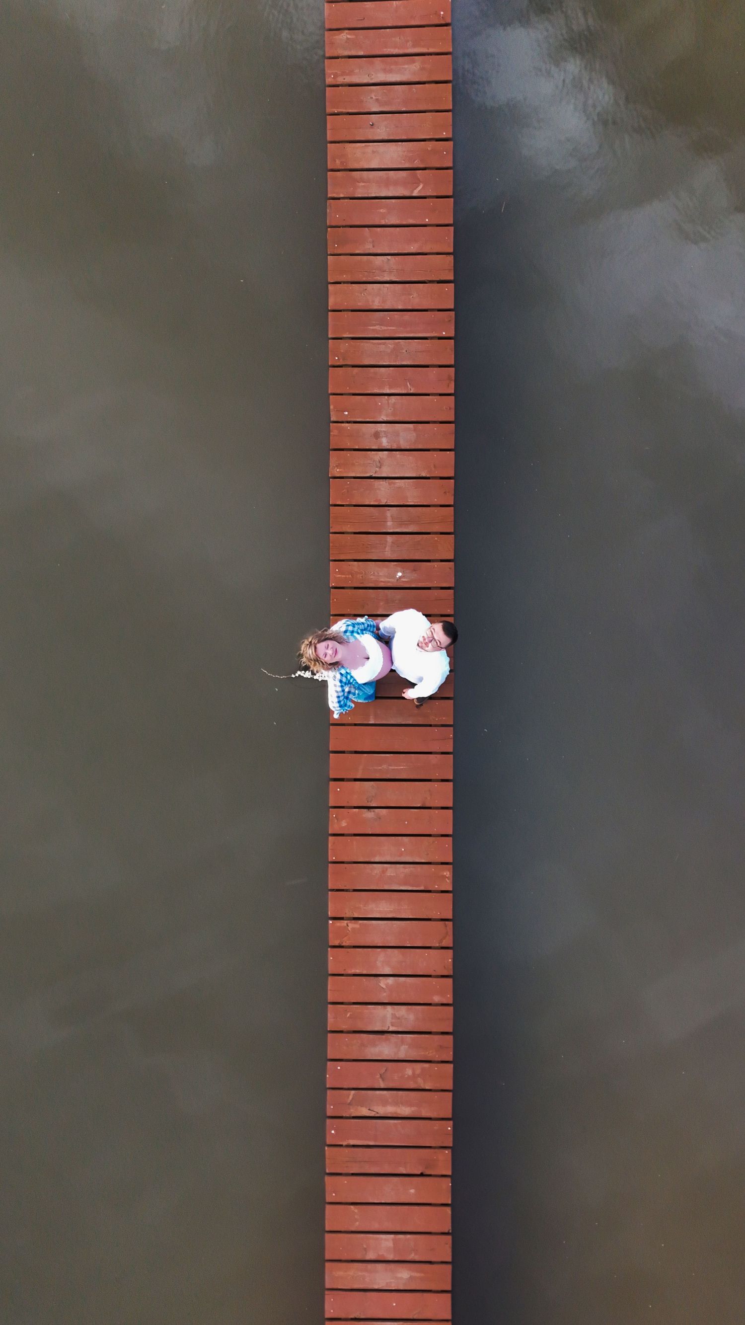 Aerial drone view of two people lying on a wooden boardwalk dock surrounded by calm water.