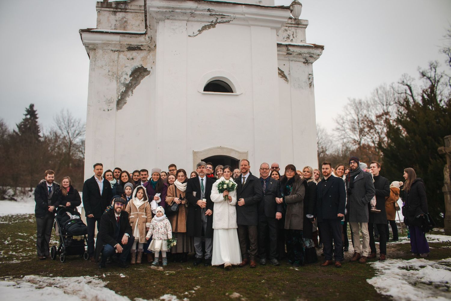 A wedding party gathers outside a white church building in winter with snow on the ground.