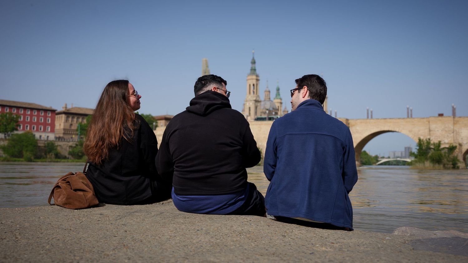 People sit along a stone wall overlooking a scenic waterfront with historic buildings and towers in the background.