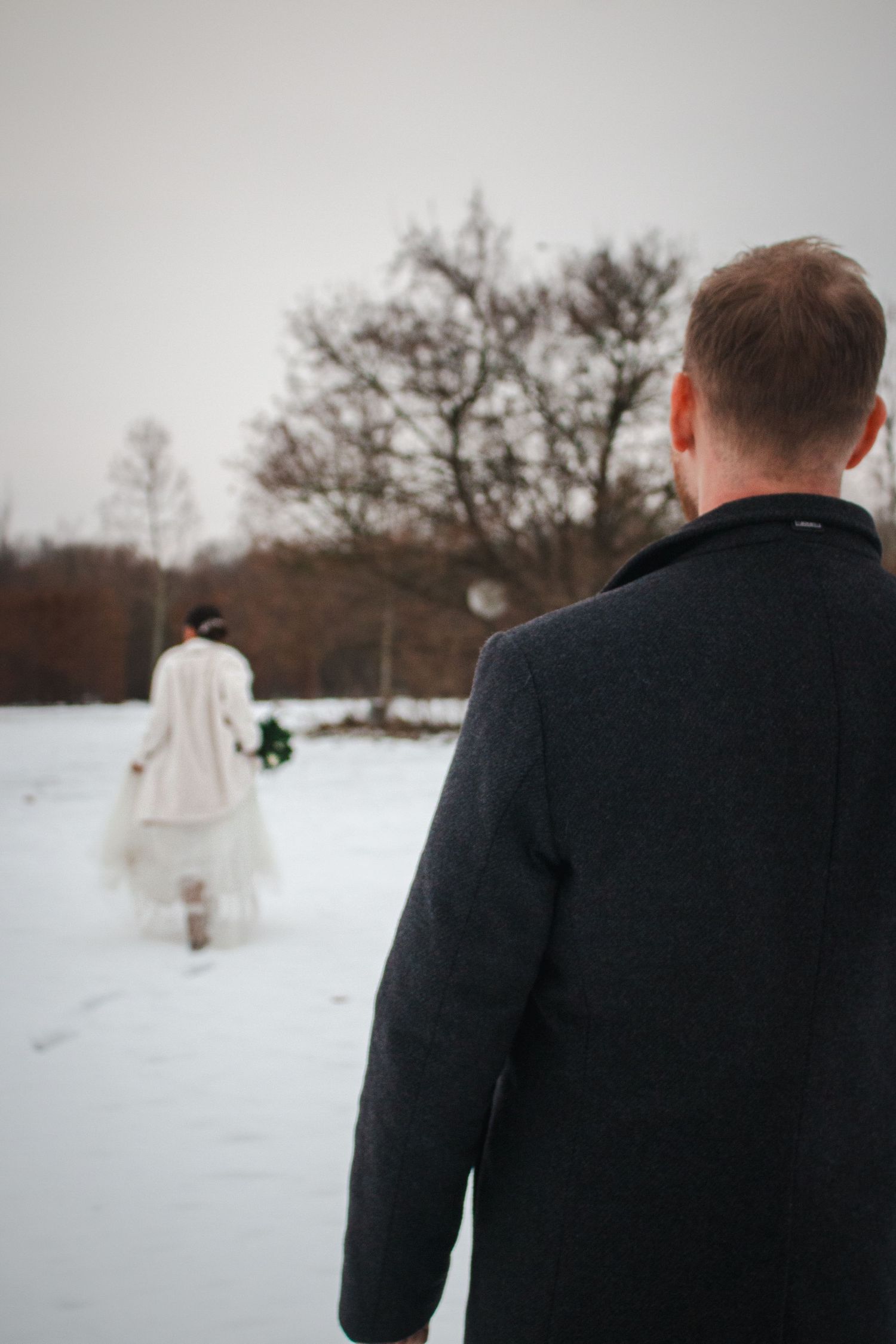 A winter wedding scene with figures walking through fresh snow surrounded by bare trees.