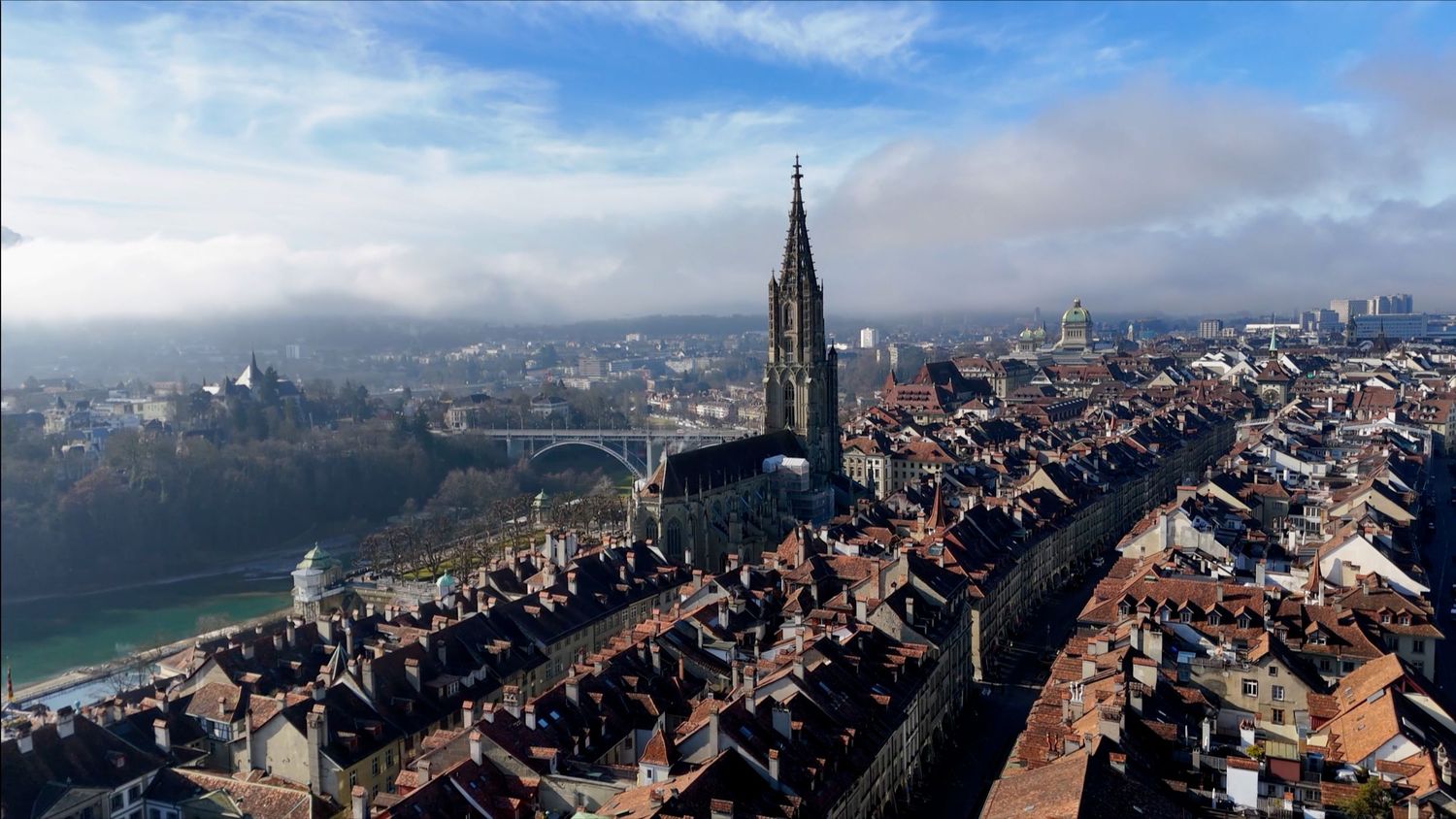 Aerial view of Bern Switzerland showing historic old town with Gothic cathedral spire and winding river under blue sky.