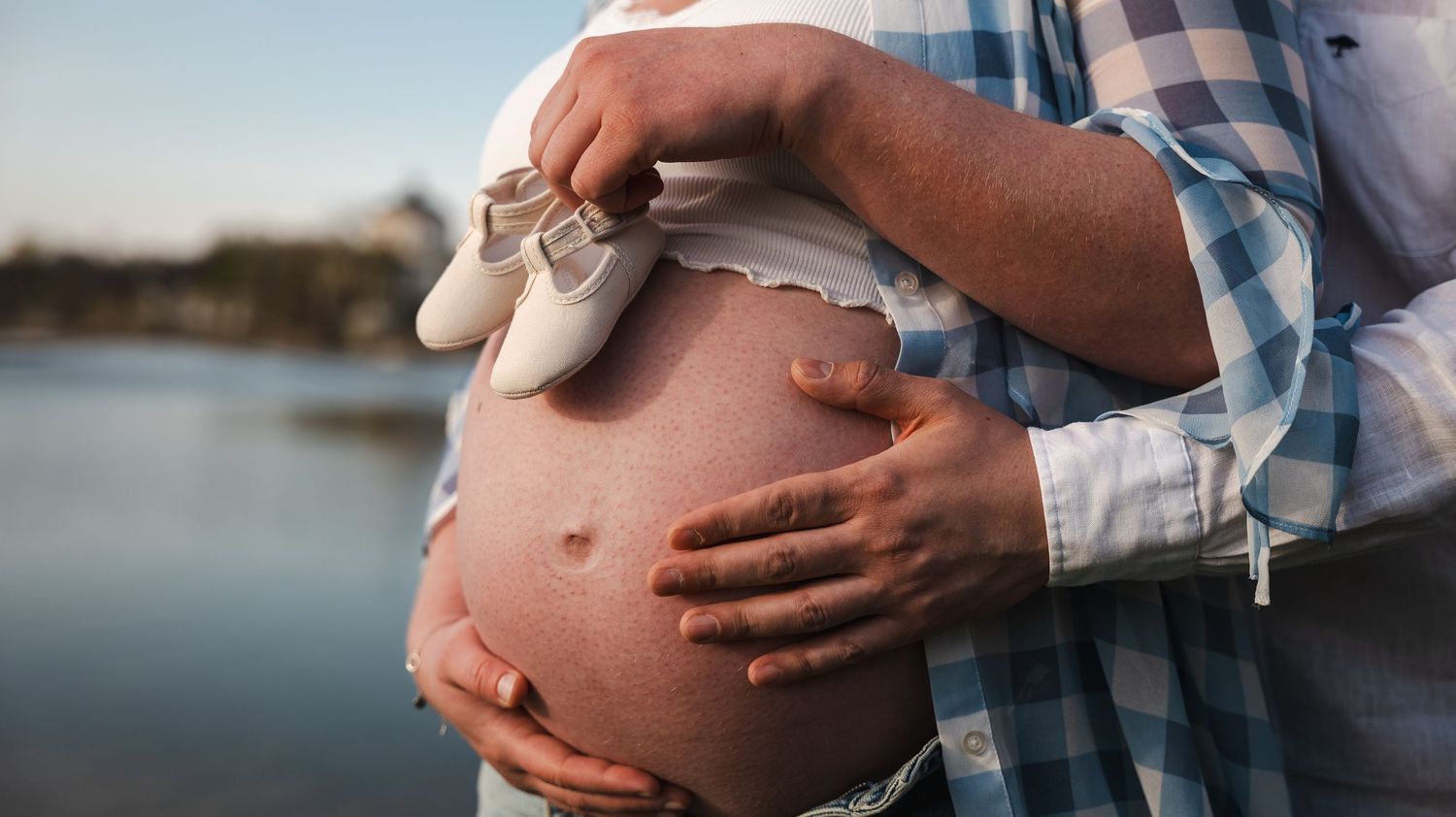Pregnant woman holding tiny baby shoes on her bare belly while partner embraces her near a serene lake.