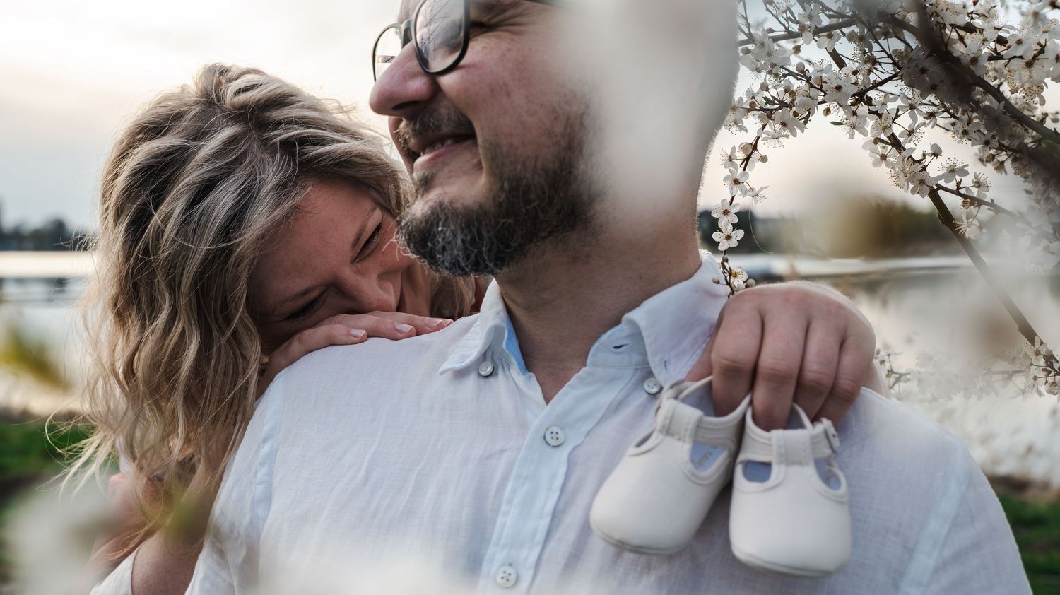 A couple embraces outdoors near blooming trees, with the man holding tiny white baby shoes, announcing their pregnancy.