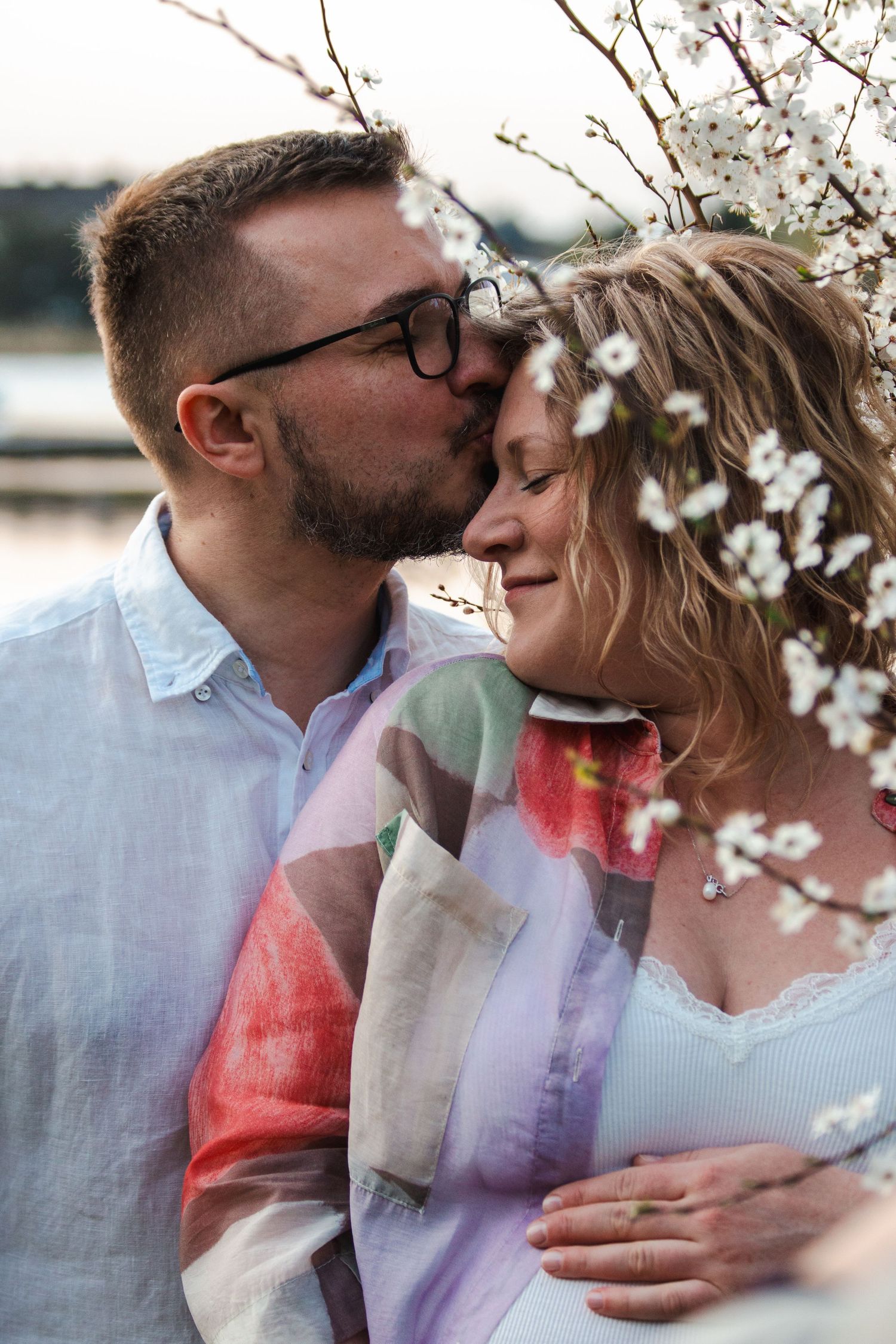 A man with glasses kisses a smiling woman near blooming white cherry blossoms in a romantic outdoor setting.