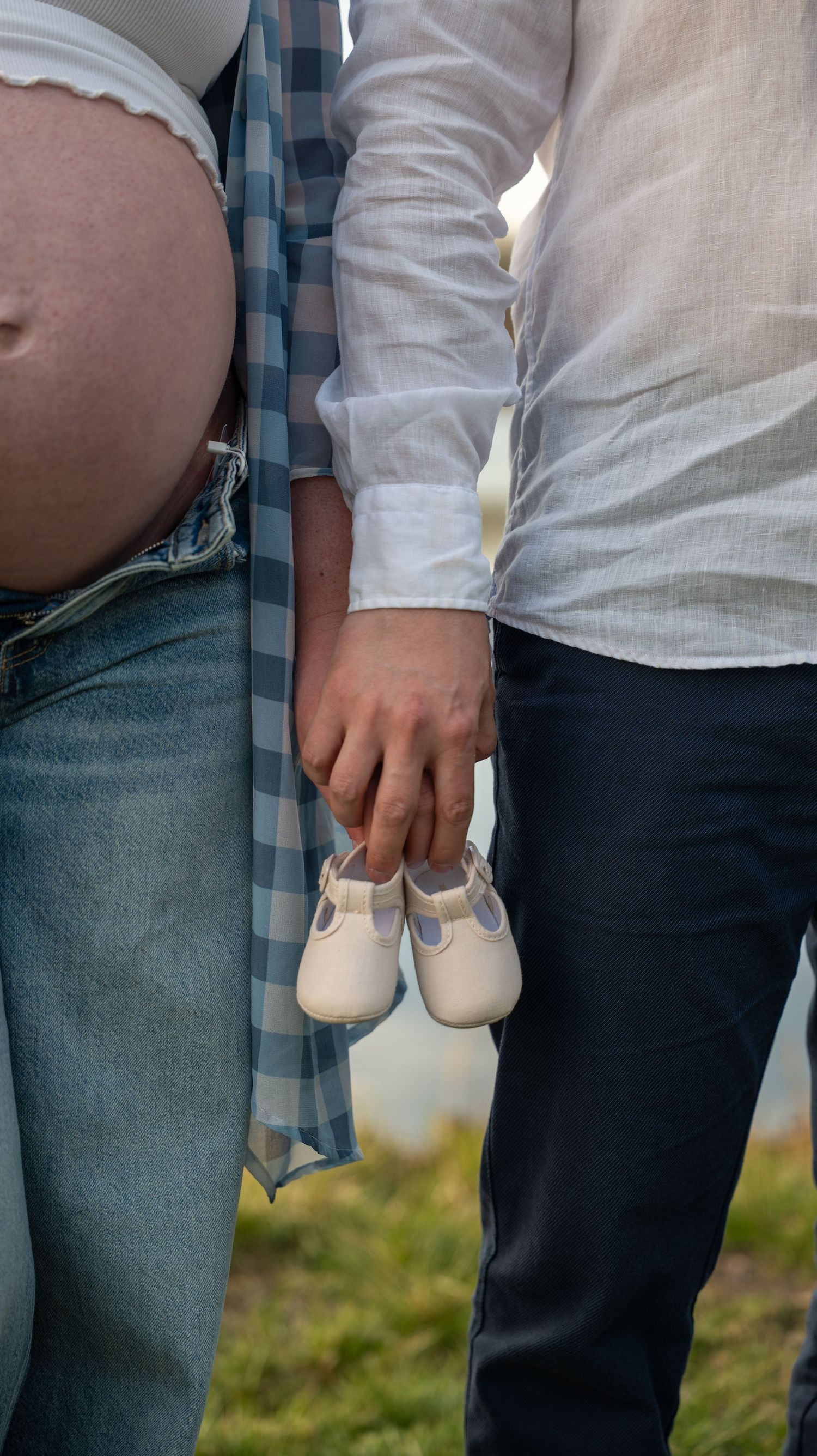 Pregnant woman and partner holding tiny white baby shoes outdoors, maternity announcement photo.