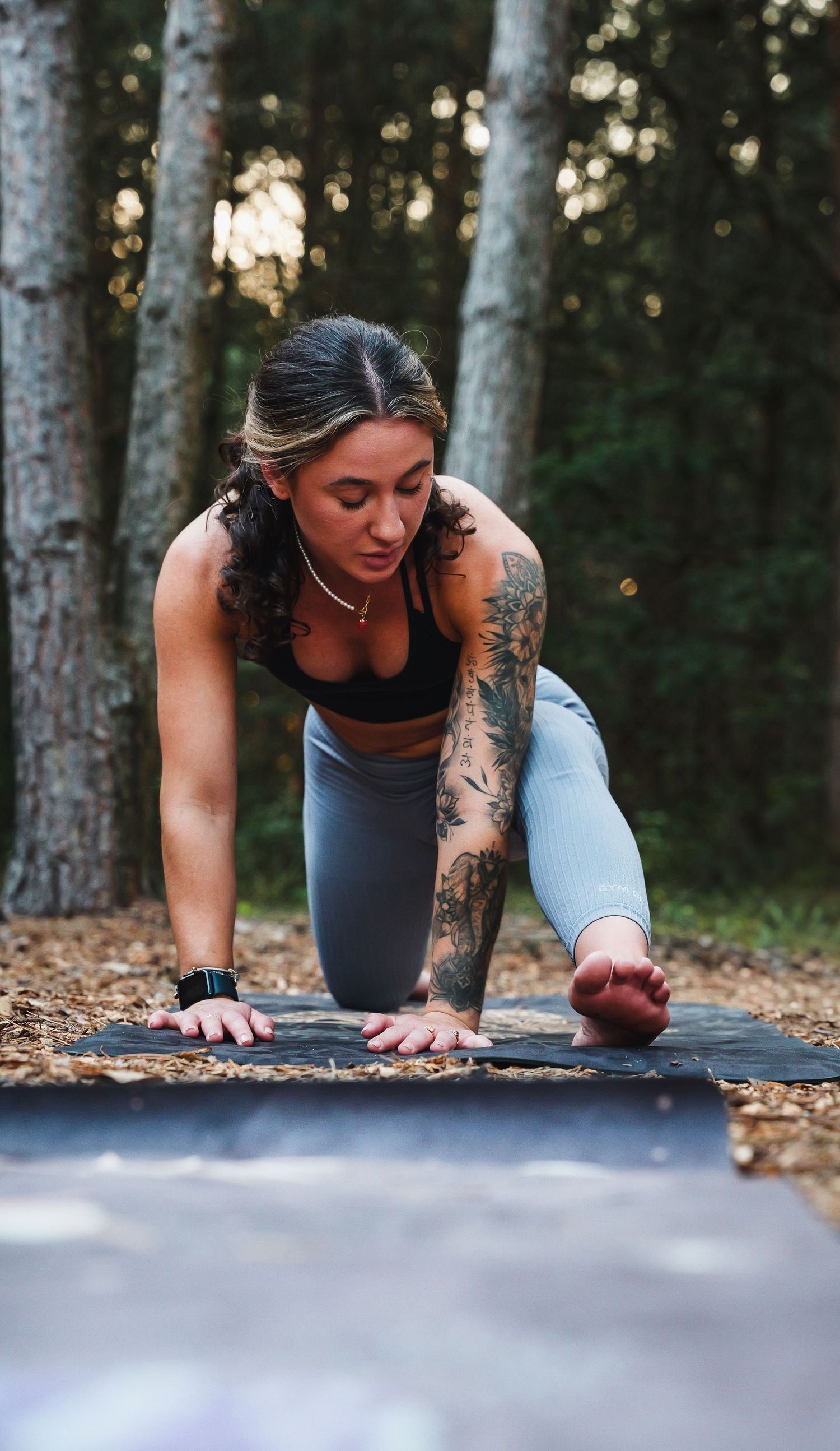 A person in workout attire performs a plank exercise on a yoga mat in a wooded outdoor setting.