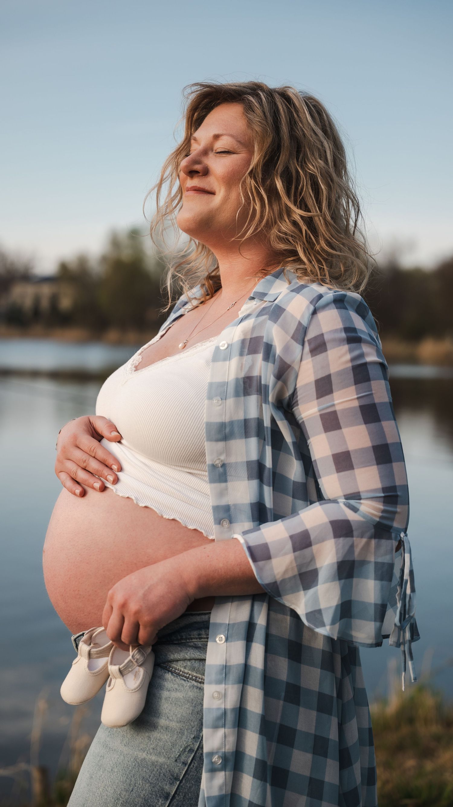 Pregnant woman holding baby shoes outdoors by a lake, cradling her belly in a plaid shirt and jeans.