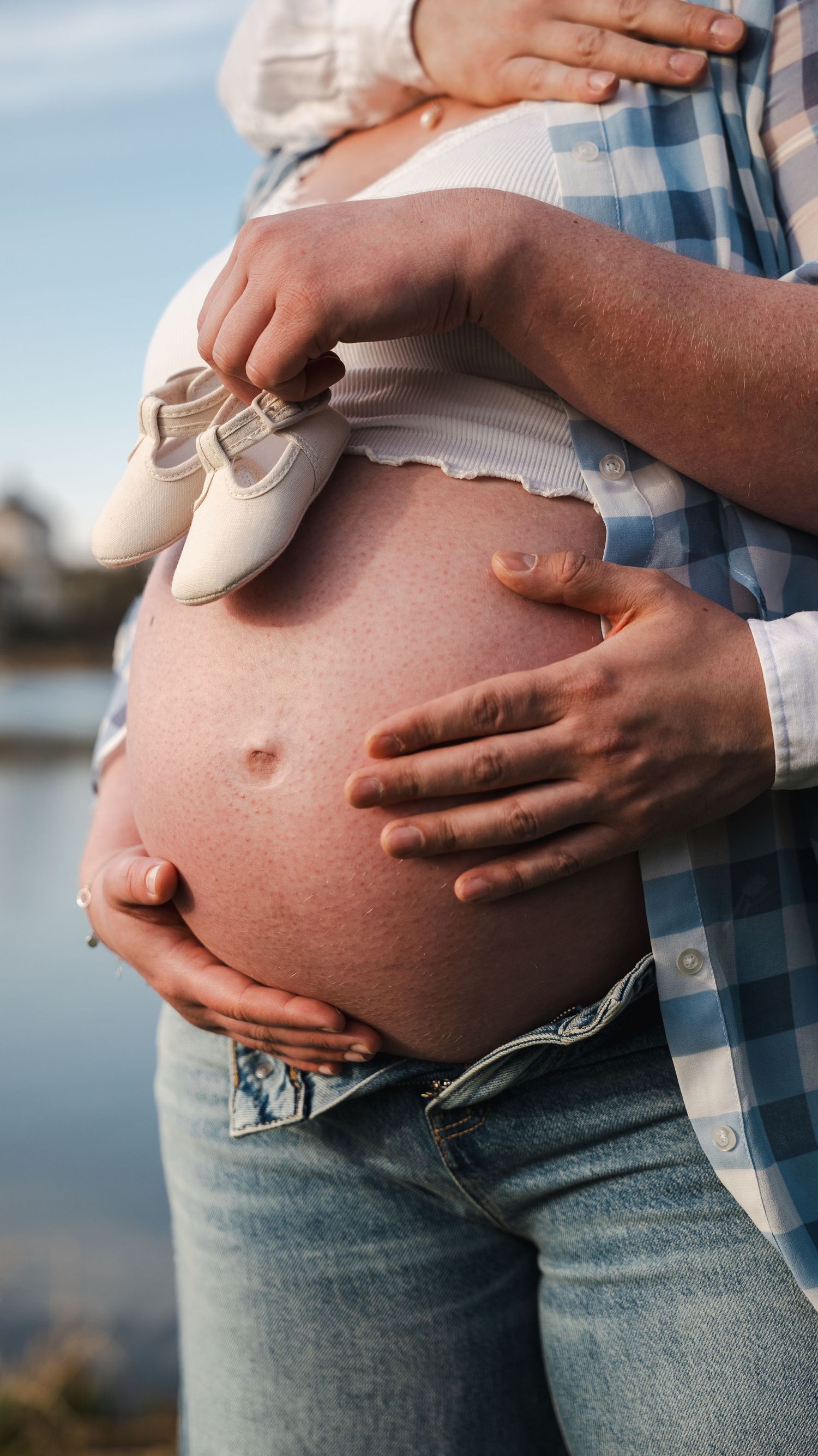 Close-up of couple holding miniature white baby shoes on pregnant belly near water.