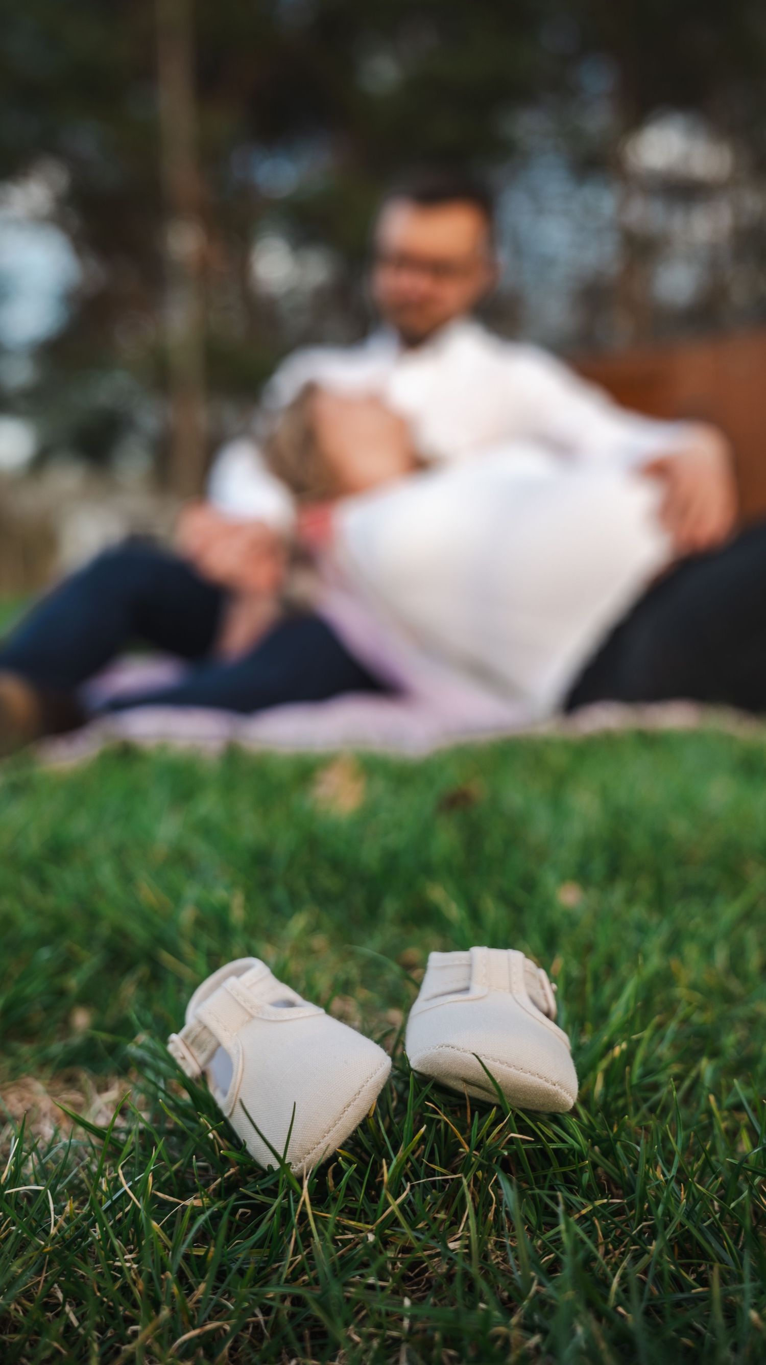 Tiny white baby shoes on green grass with blurred expectant couple sitting together in park background.