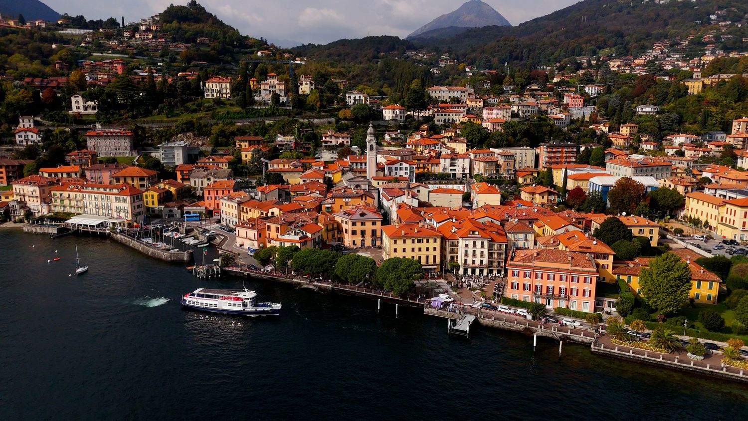 Panoramic view of a Mediterranean coastal town with red-roofed buildings nestled against mountainous terrain and harbor.